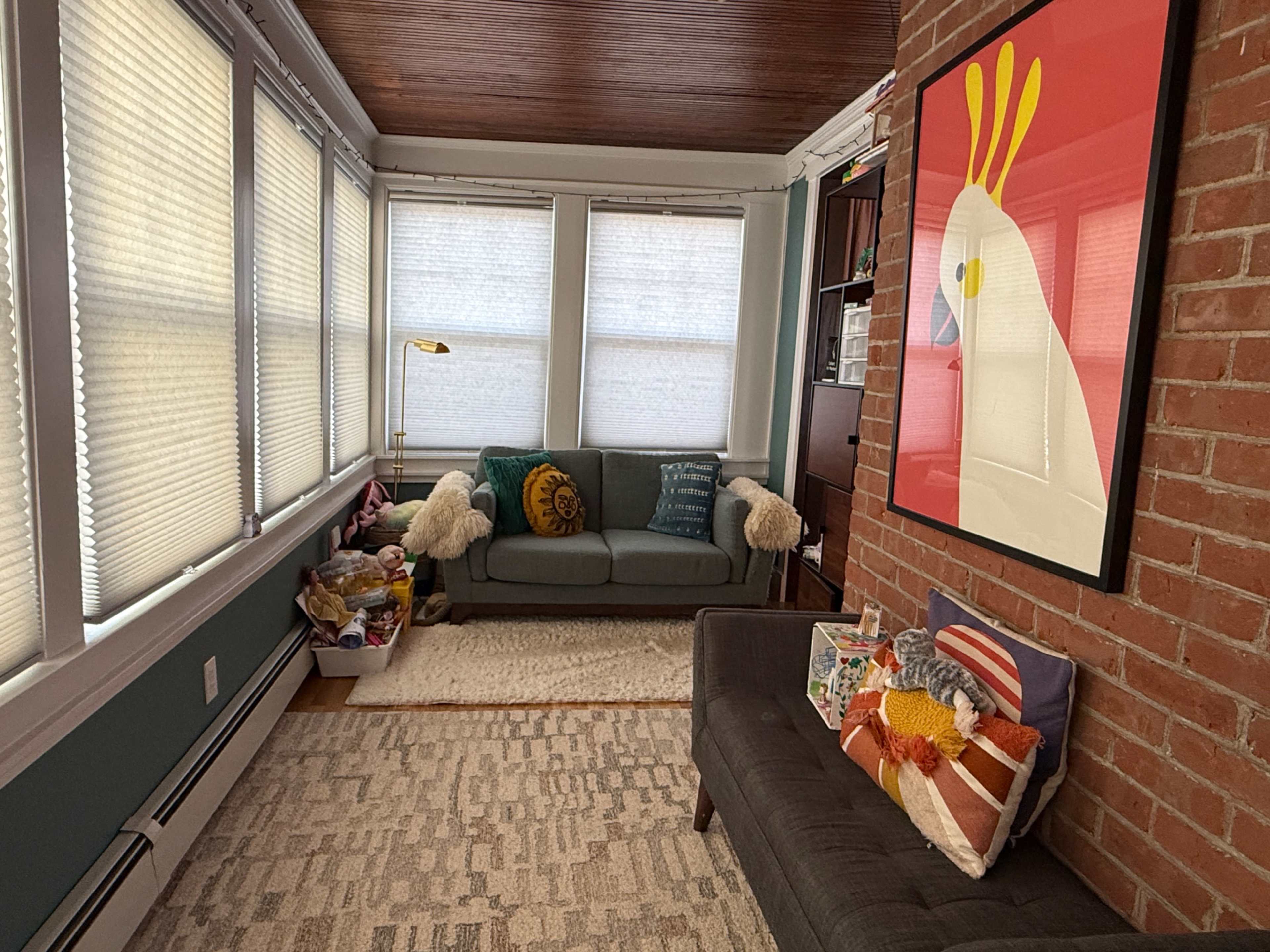 The image shows a cozy living space with a gray sofa, a dark gray couch, a large colorful artwork of a chicken on the wall, and large windows covered with shades, along with a textured rug on the floor.