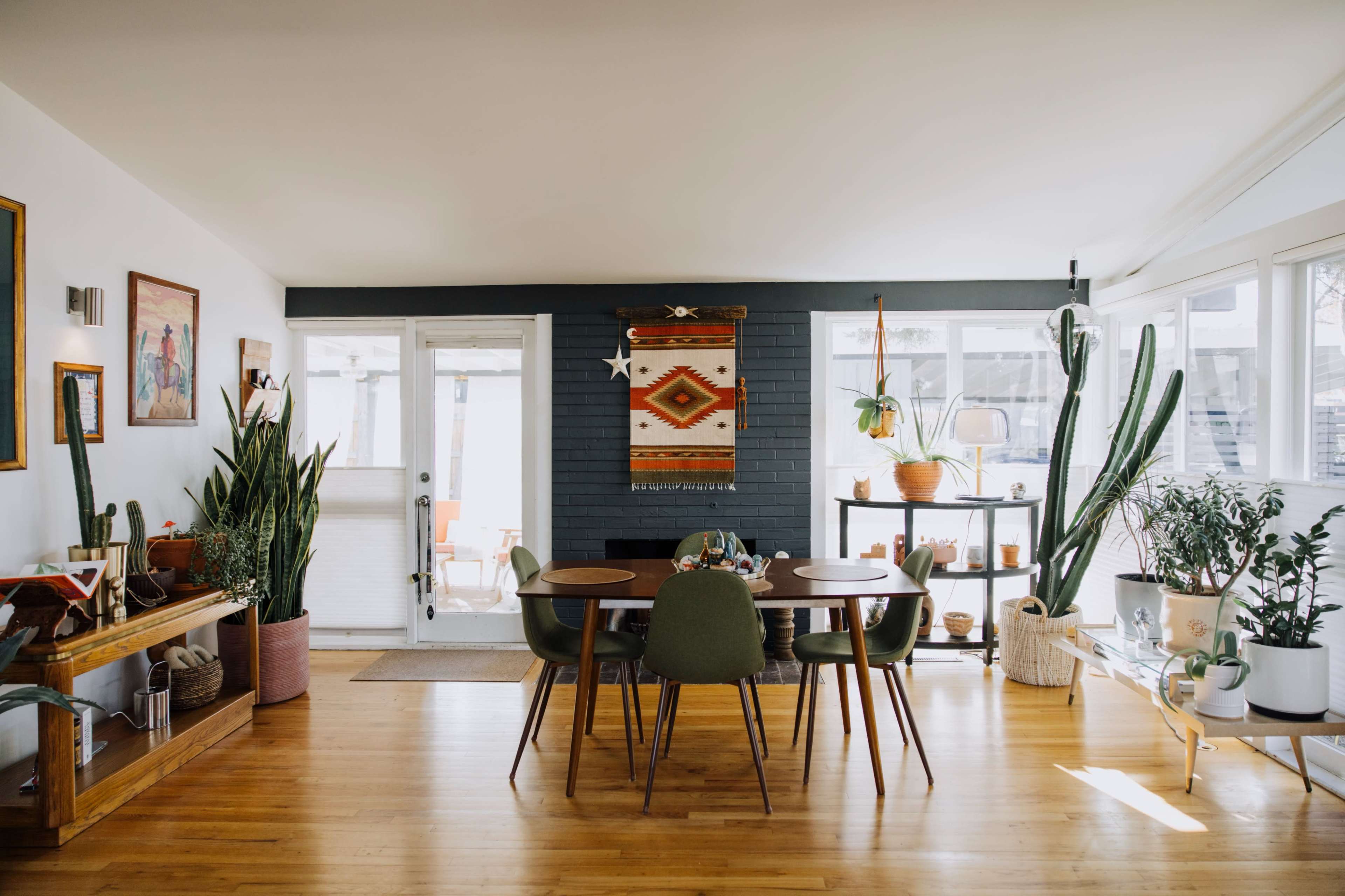 The image shows a modern dining area with a wooden table surrounded by green chairs, indoor plants, and a decorative wall hanging.