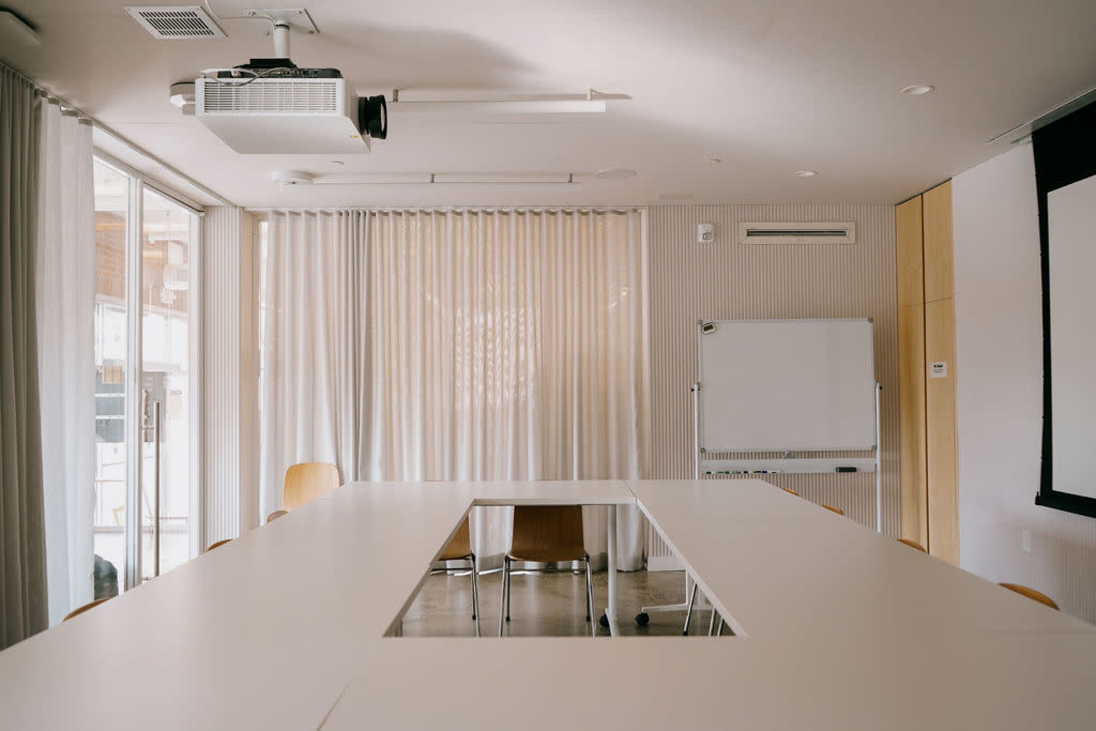 A rectangular conference table with a projector and whiteboard is set up in a well-lit meeting room with floor-to-ceiling windows and curtains.