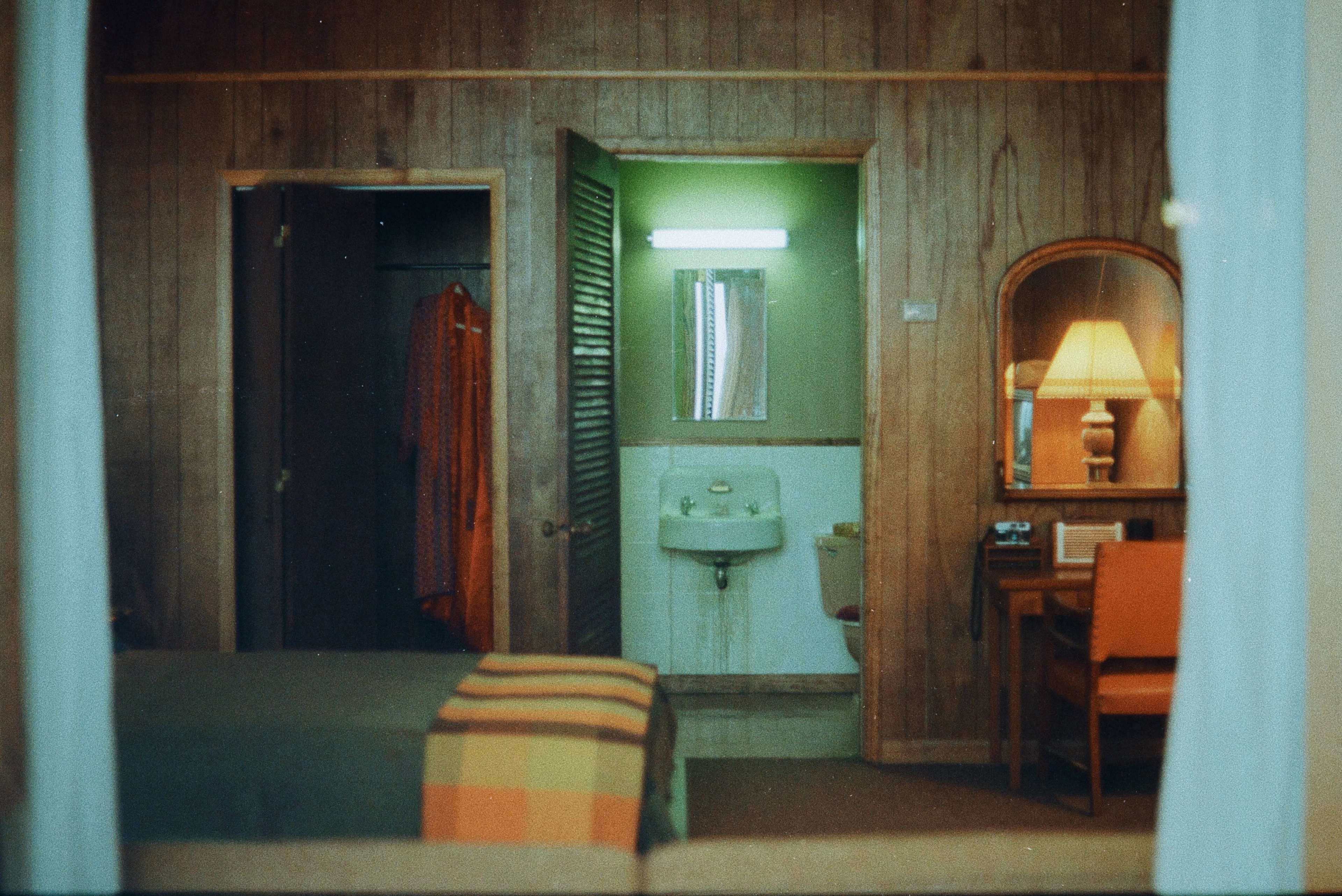 The image shows a rustic hotel room with a bed, a mirror, a lamp, and a bathroom visible through an open door.