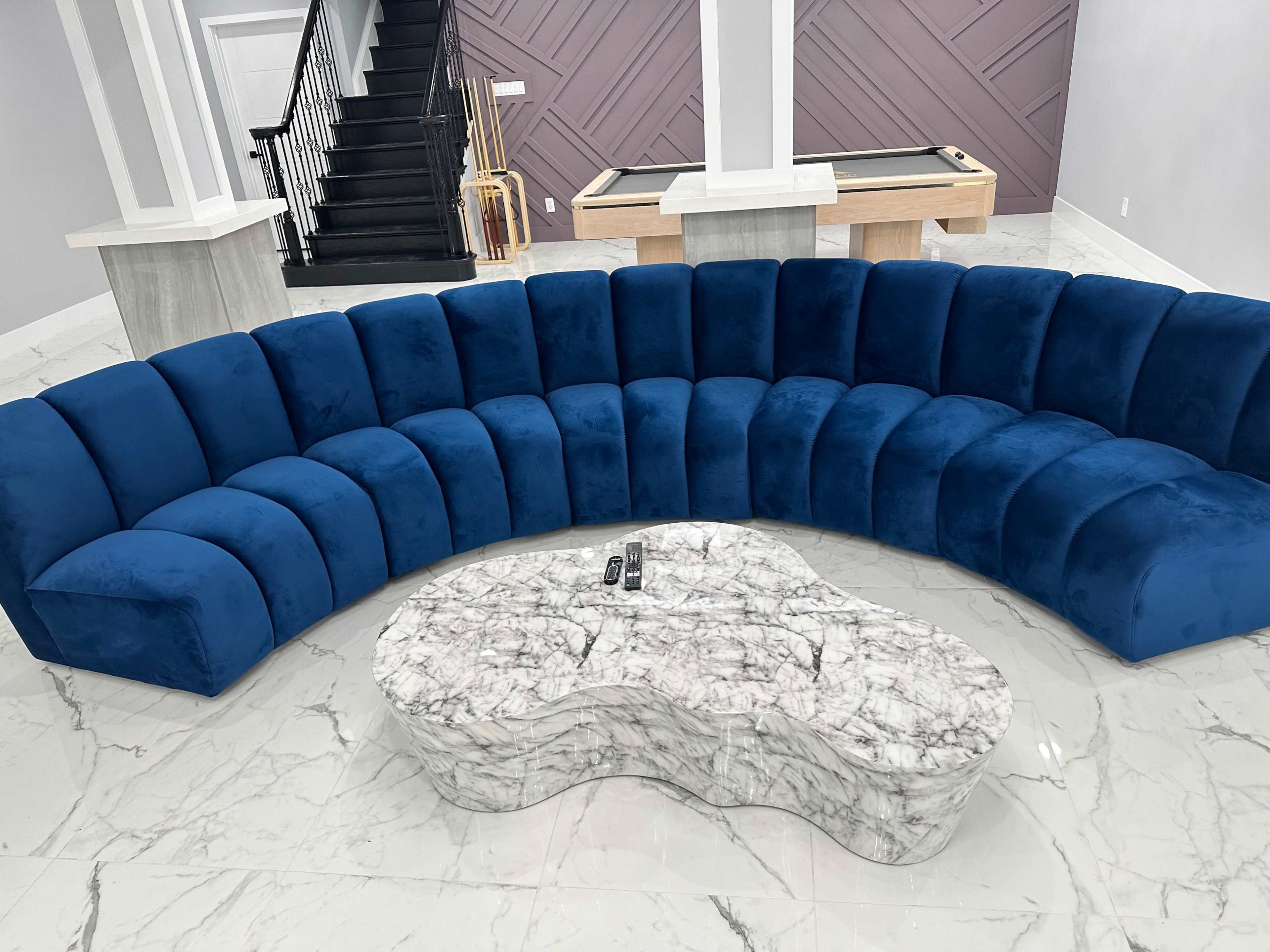 A curved blue velvet sofa arranged in a semi-circle around a marble coffee table in a spacious indoor area with a staircase in the background.
