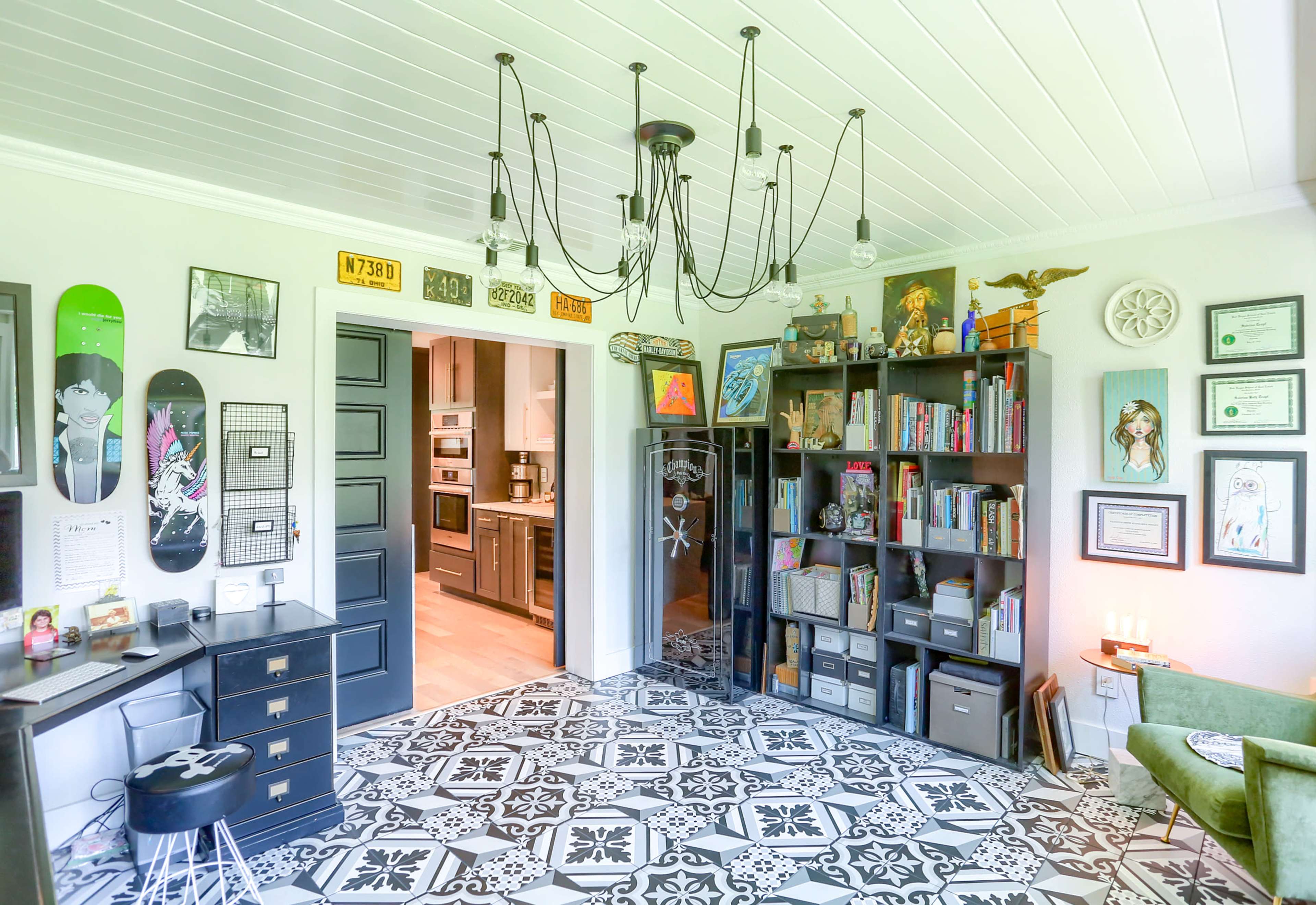 The room features a black bookshelf filled with books and decor, a workspace with a desk, a patterned tiled floor, and an adjoining doorway leading to a kitchen area.