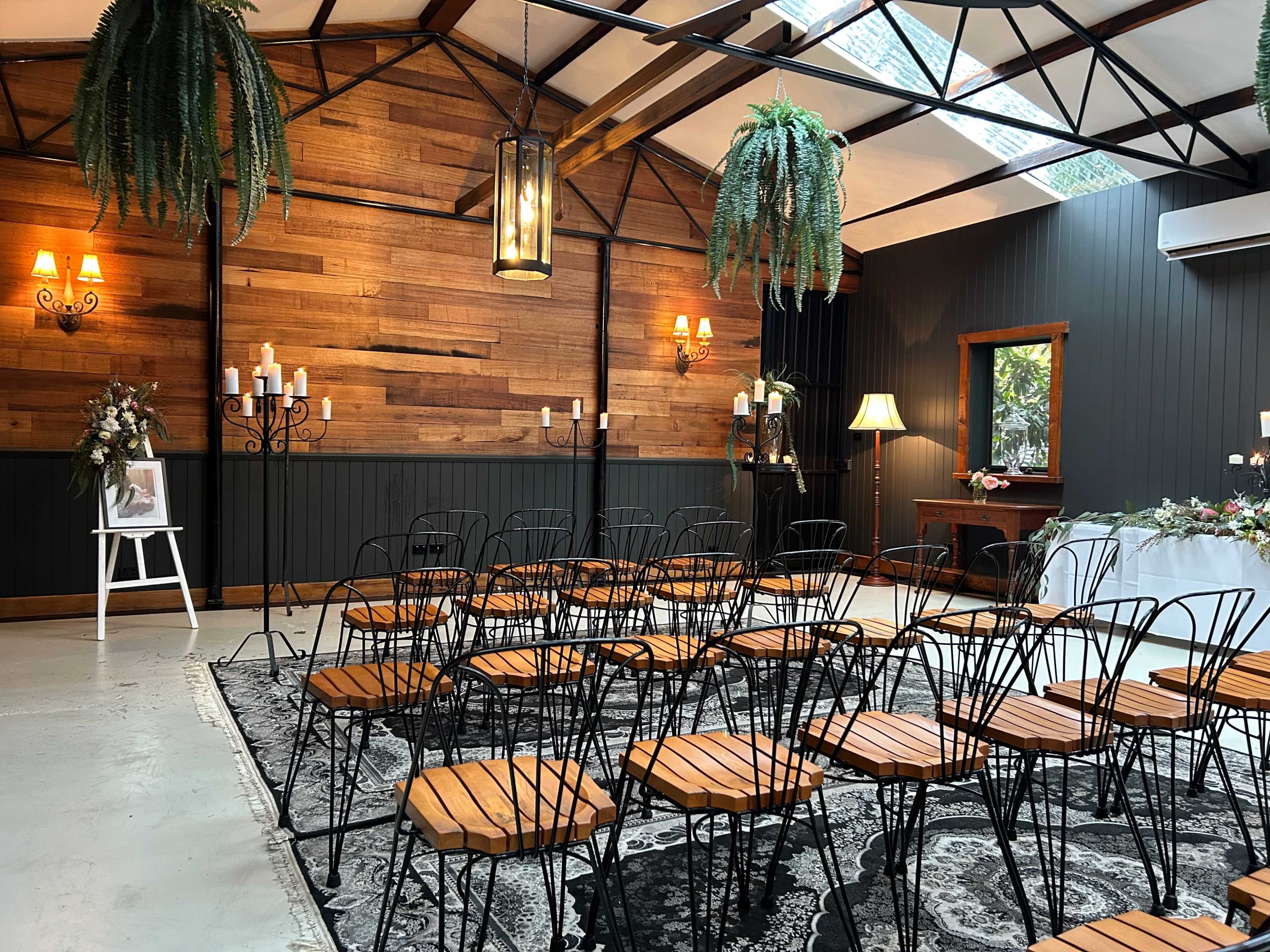 A spacious indoor venue features rows of black metal chairs with wooden seats, surrounded by wood-paneled walls and decorative lighting.