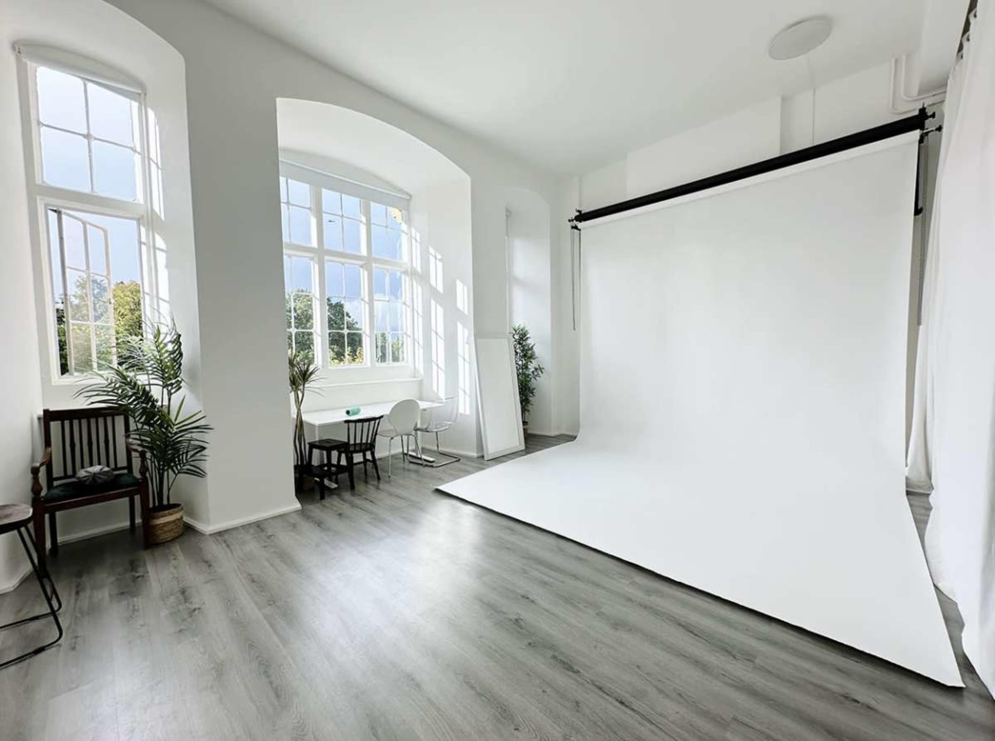 The image shows a bright, spacious photo studio with large windows, a white backdrop, and minimal furniture.