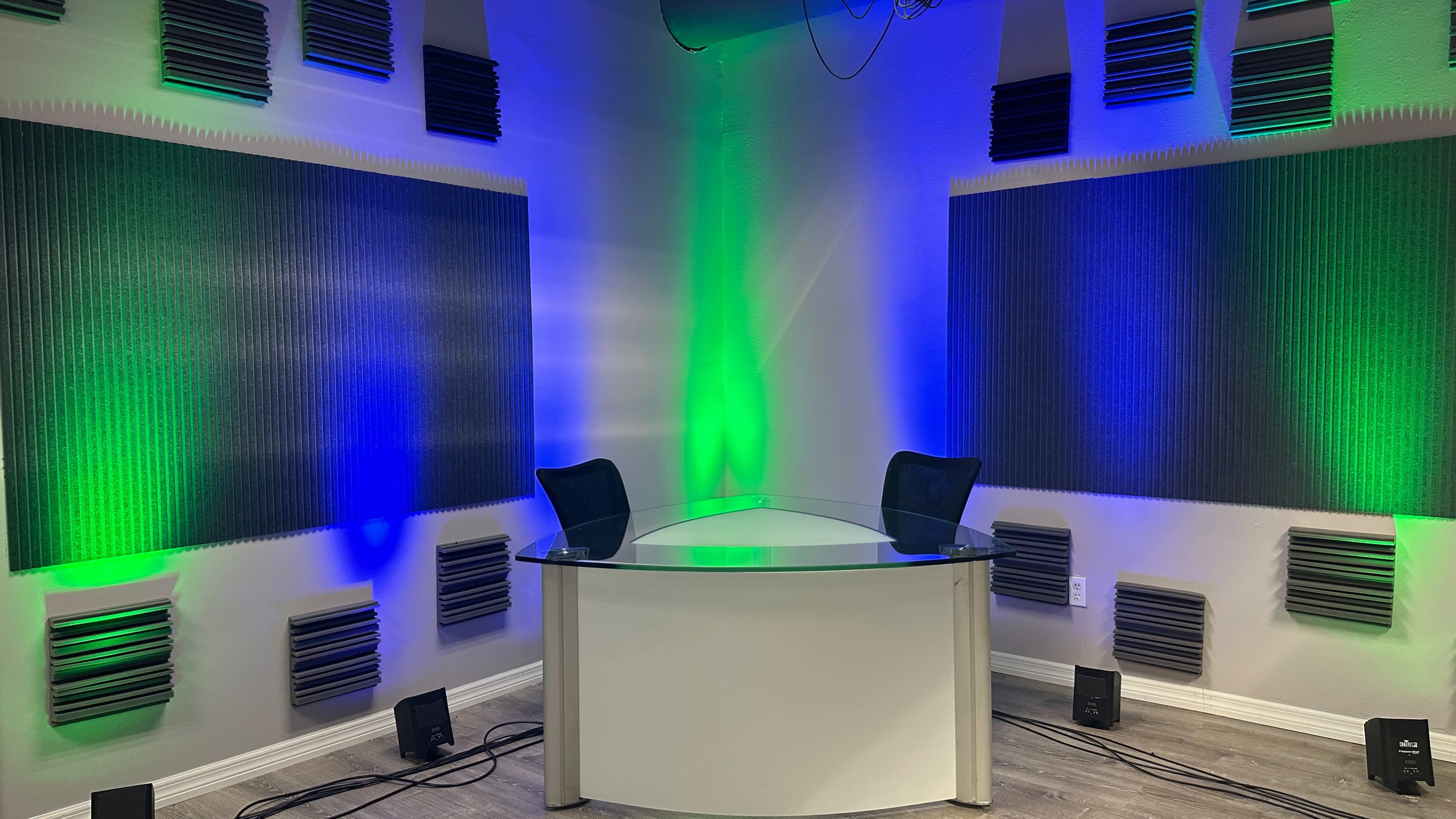 A modern, minimalistic studio with a glass desk and two black chairs, surrounded by wall-mounted acoustic panels and illuminated by green and blue lighting.