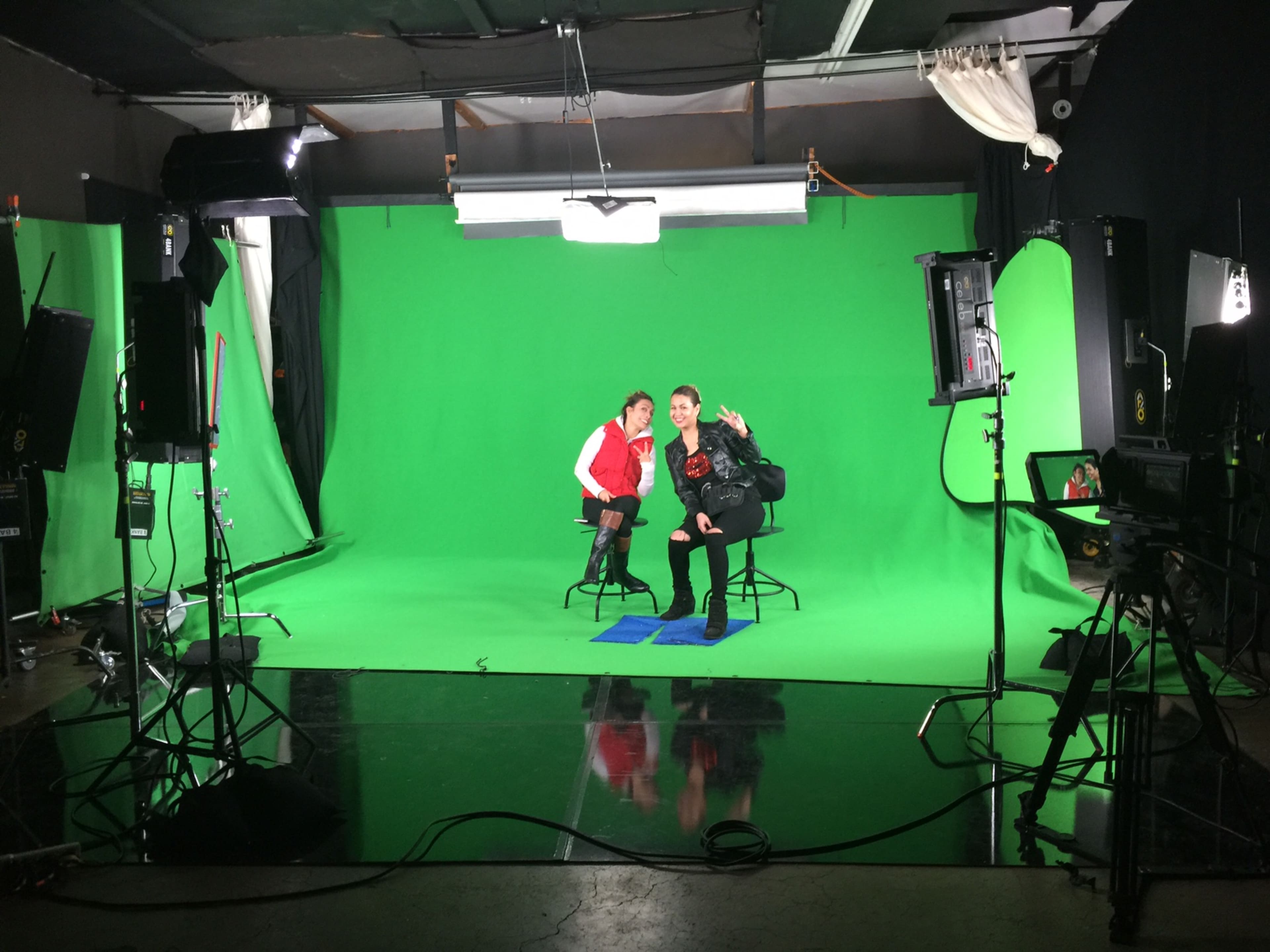 Two individuals are seated on high stools in a studio with a green screen backdrop, surrounded by lighting equipment.