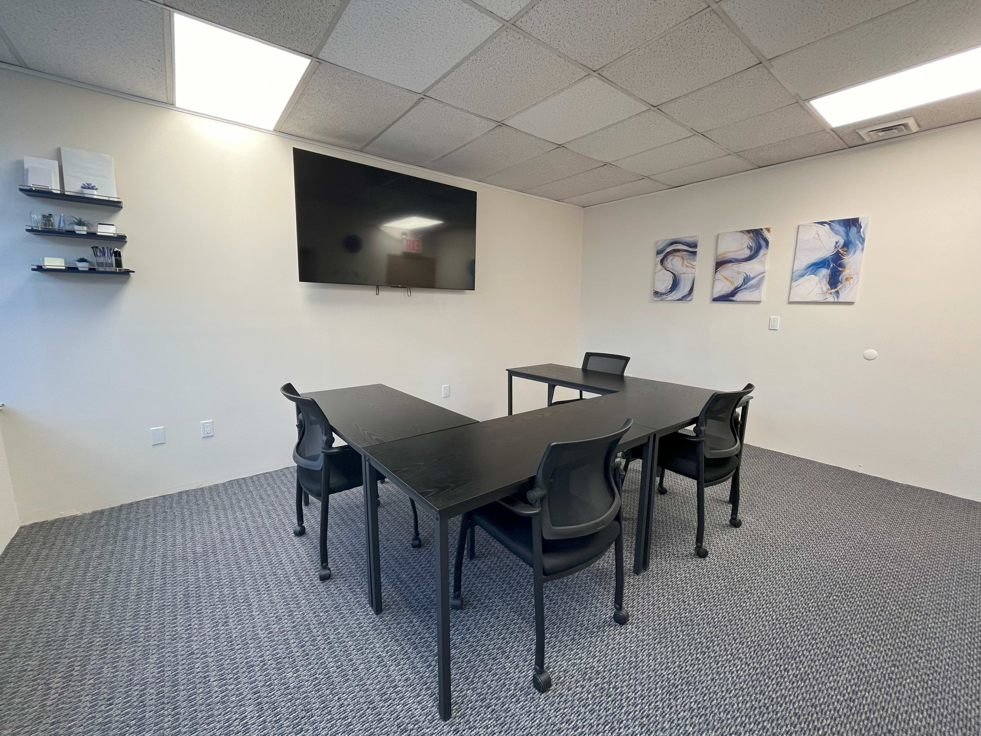 A simple meeting room features a large television mounted on the wall, four black chairs, and abstract artwork.
