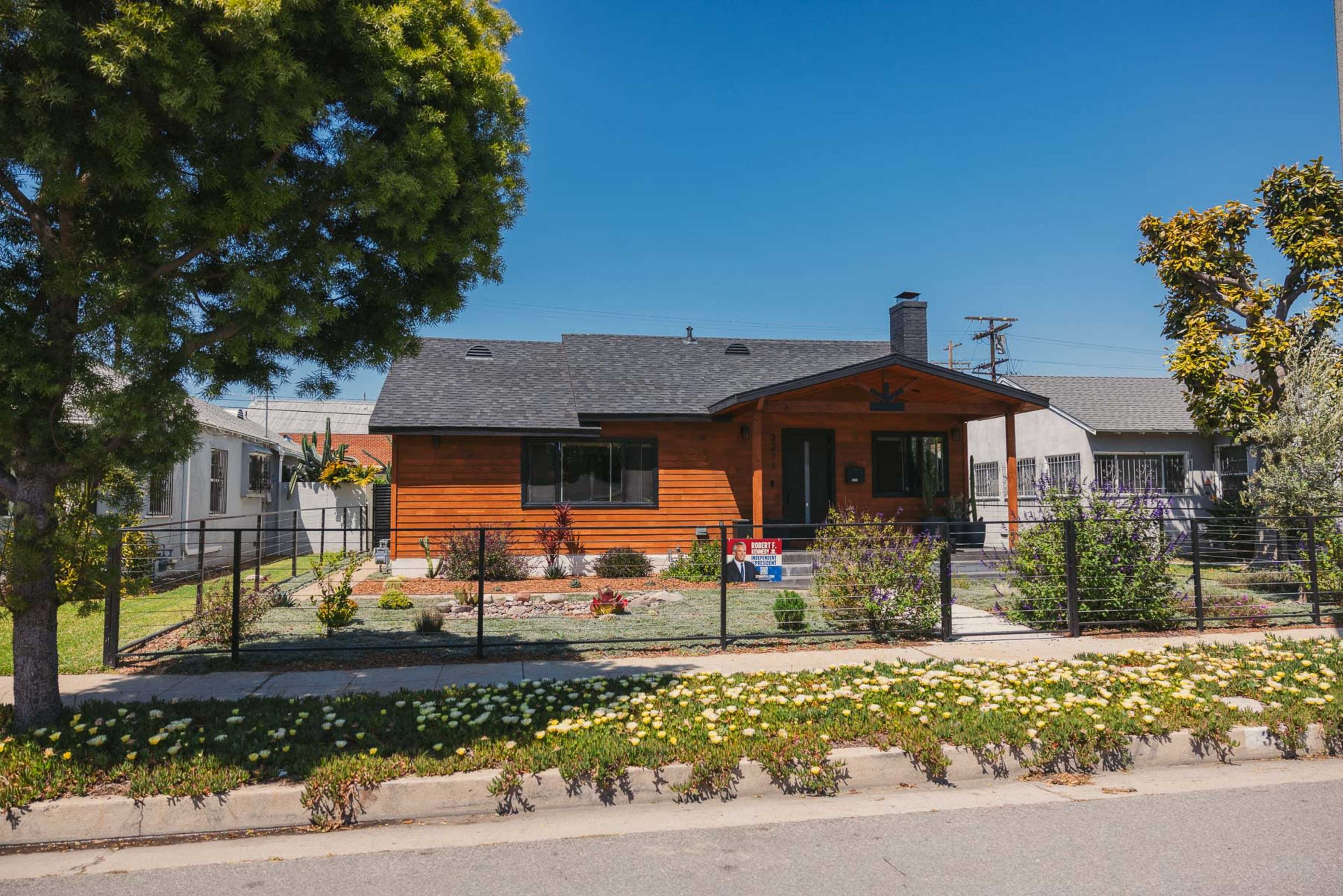 The image features a single-story wooden house with a black roof, surrounded by a garden and a black fence along the sidewalk.