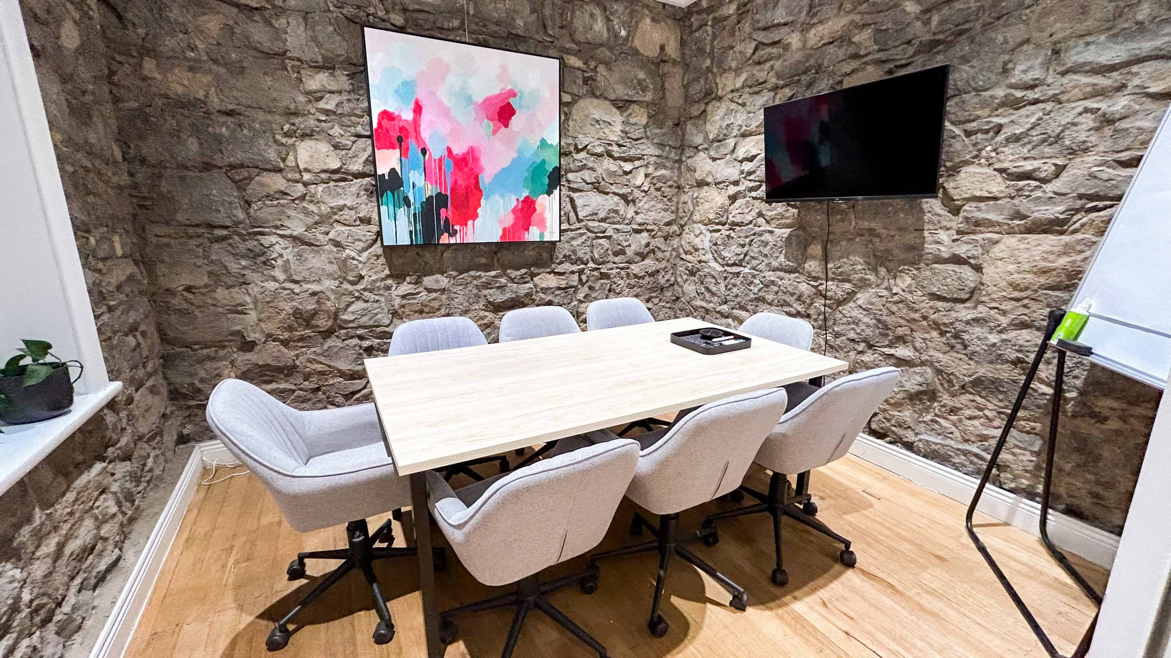 A conference room features a wooden table surrounded by gray chairs, with a colorful abstract painting on the stone wall and a wall-mounted TV.