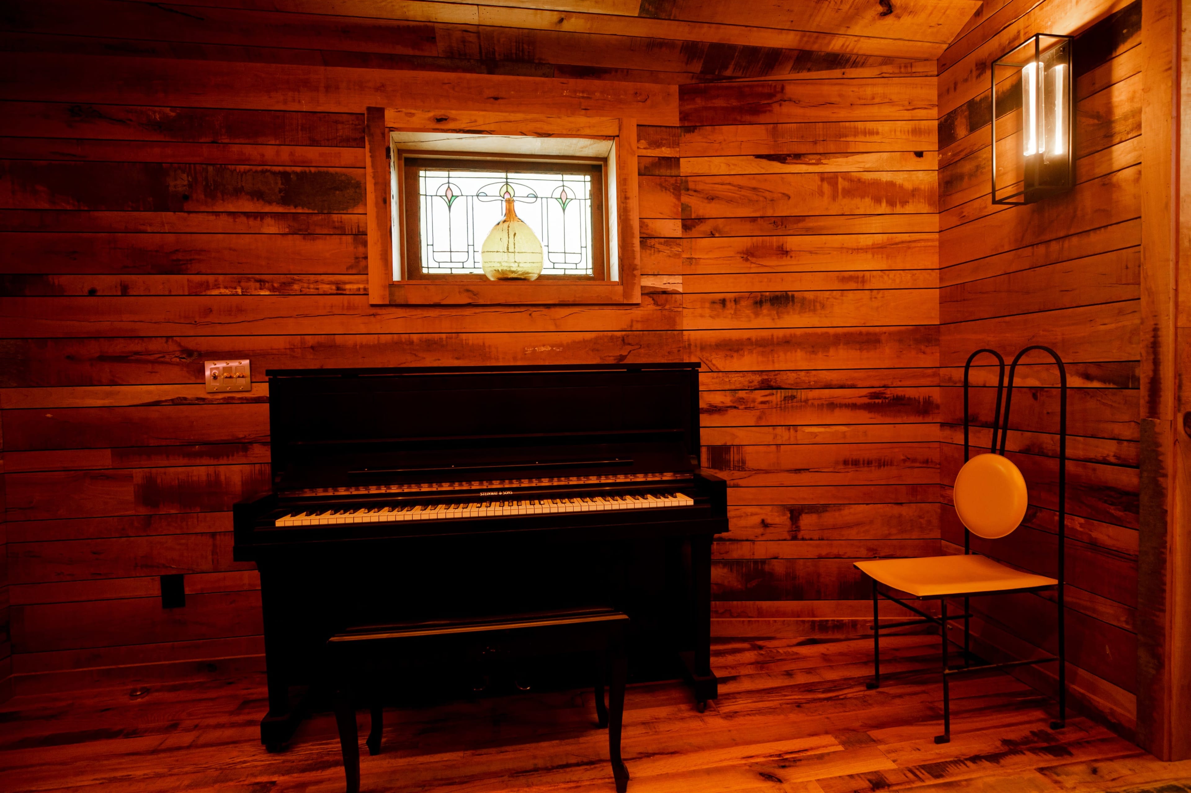 A black piano sits against a wooden wall in a warmly lit room with a window featuring a stained glass design.
