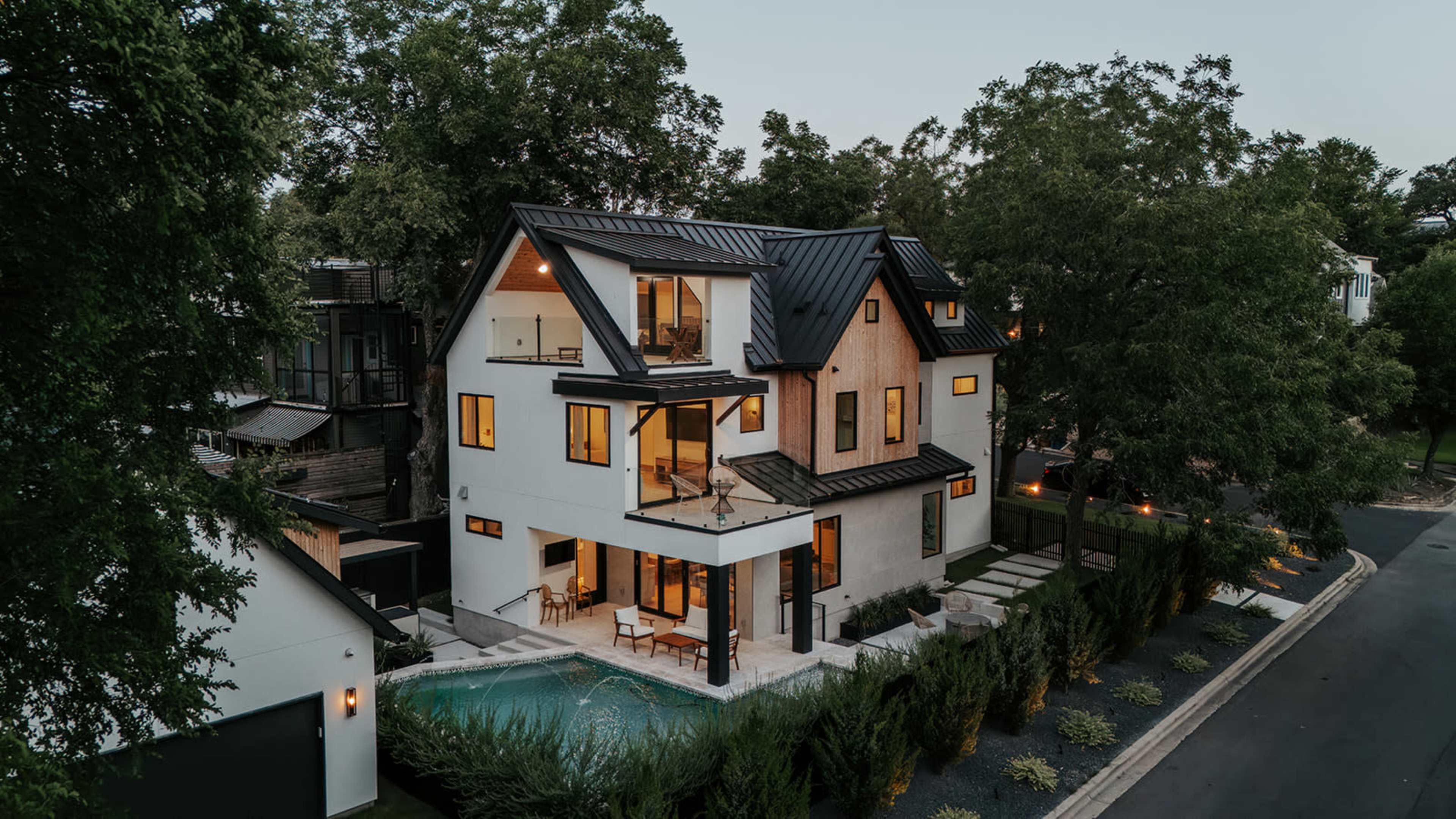 A modern two-story house with a black metal roof features large windows, a swimming pool, and a patio surrounded by lush greenery.