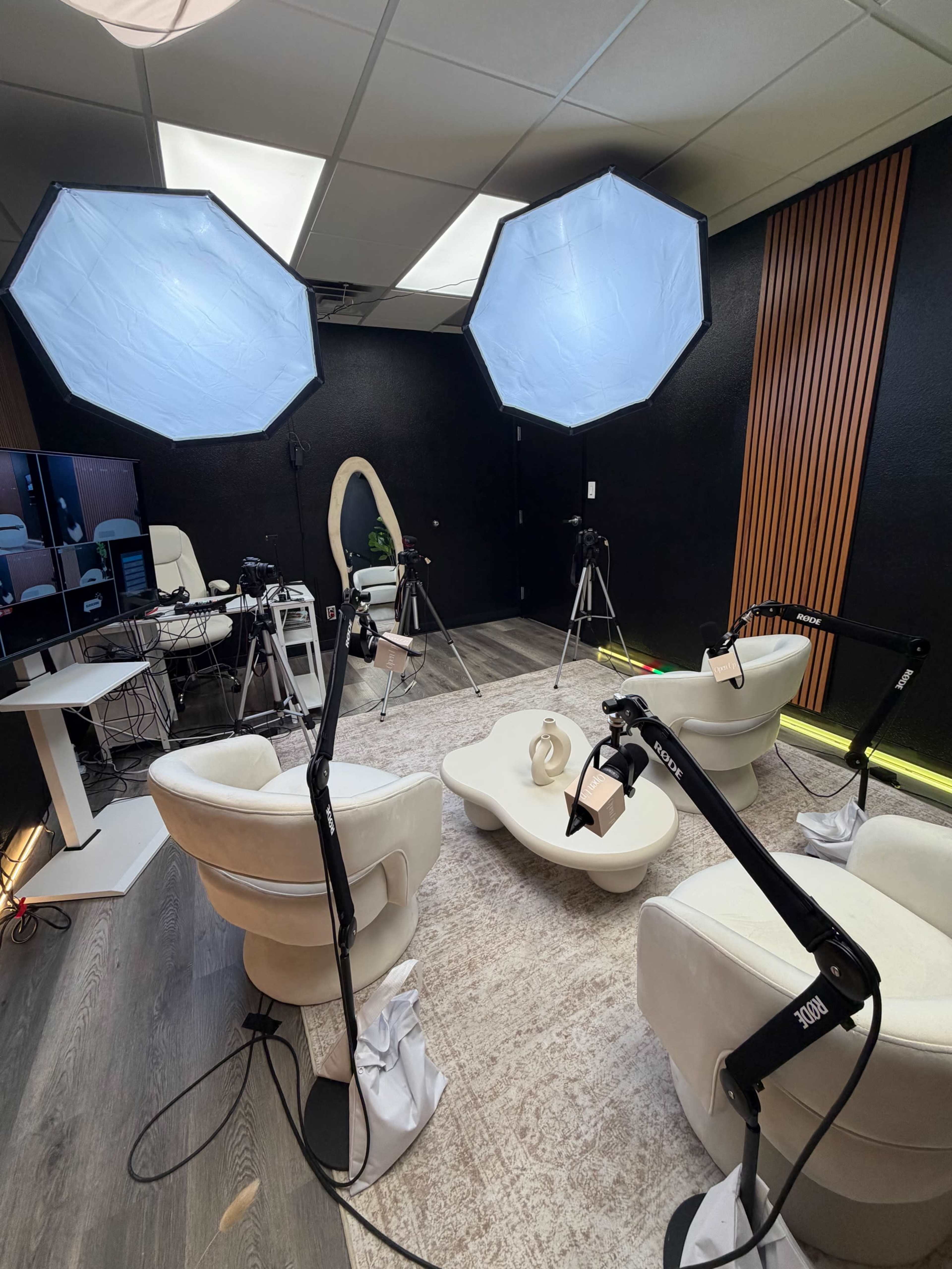 The image shows a well-lit studio setup featuring two softbox lights, two camera tripods, and two white armchairs arranged around a decorative table on a carpeted floor.