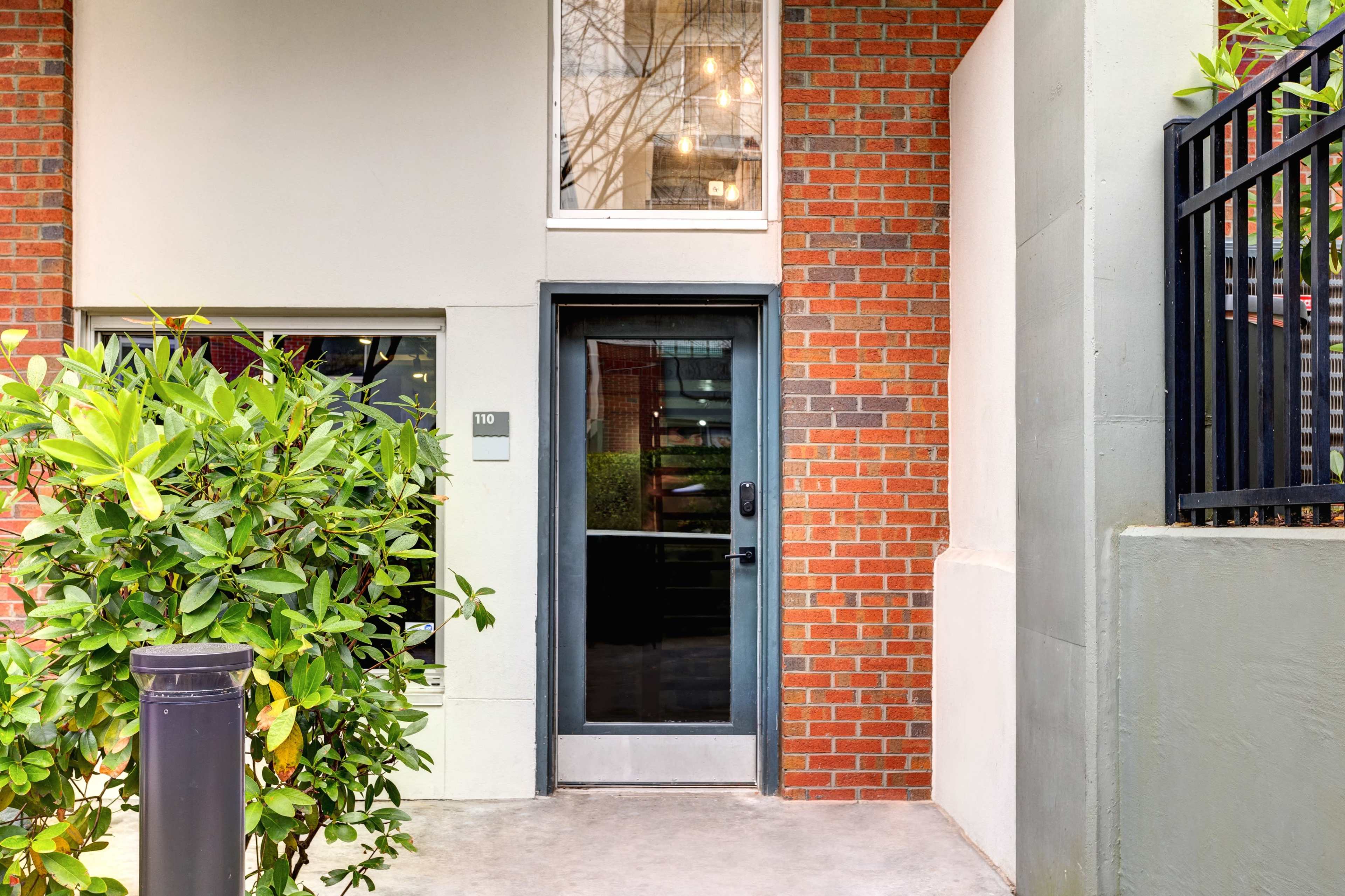 A modern entrance with a black door framed by brick walls and greenery on the side.