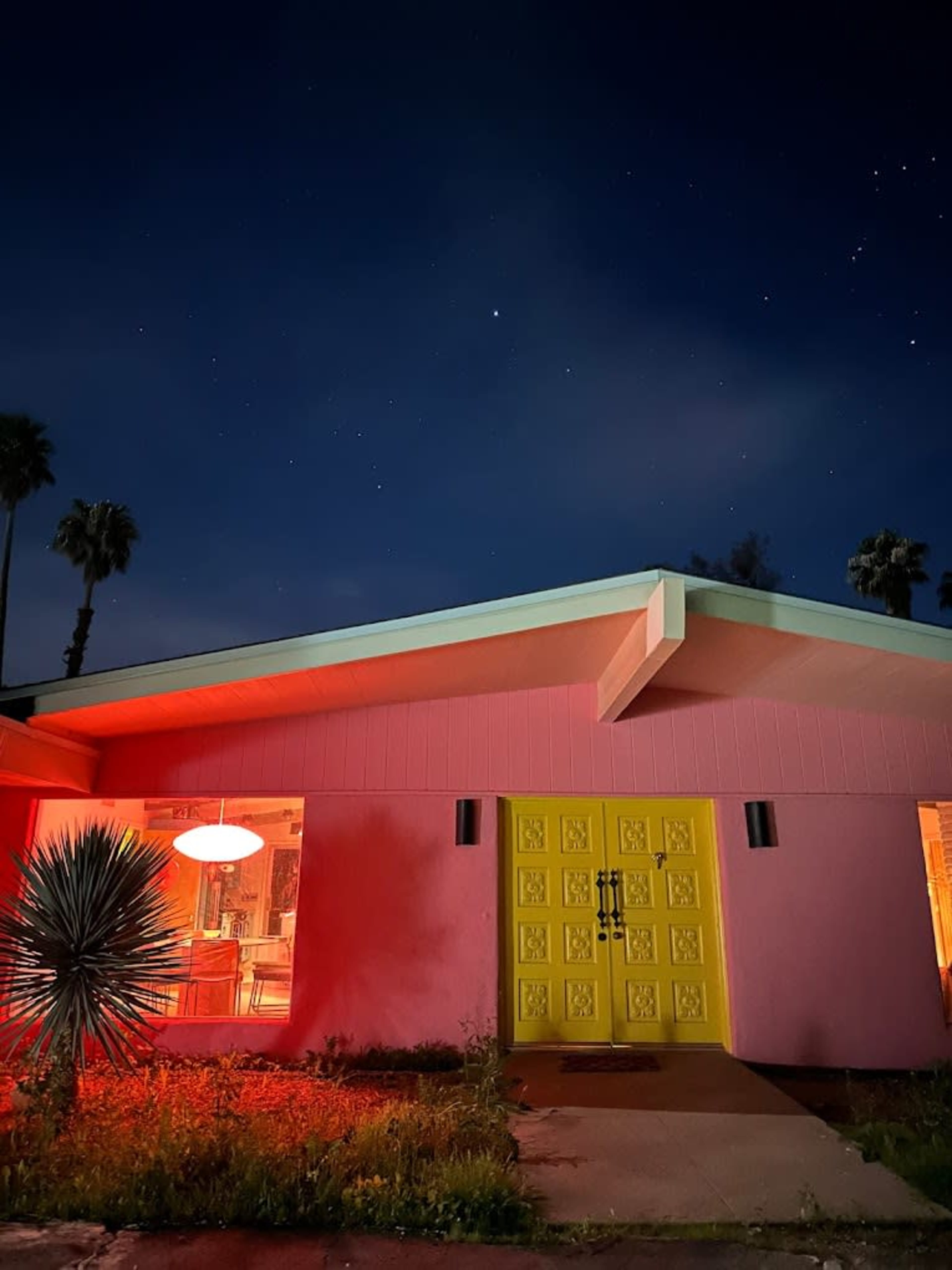 A pink house with a bright yellow door is illuminated by red light at night, under a starry sky.