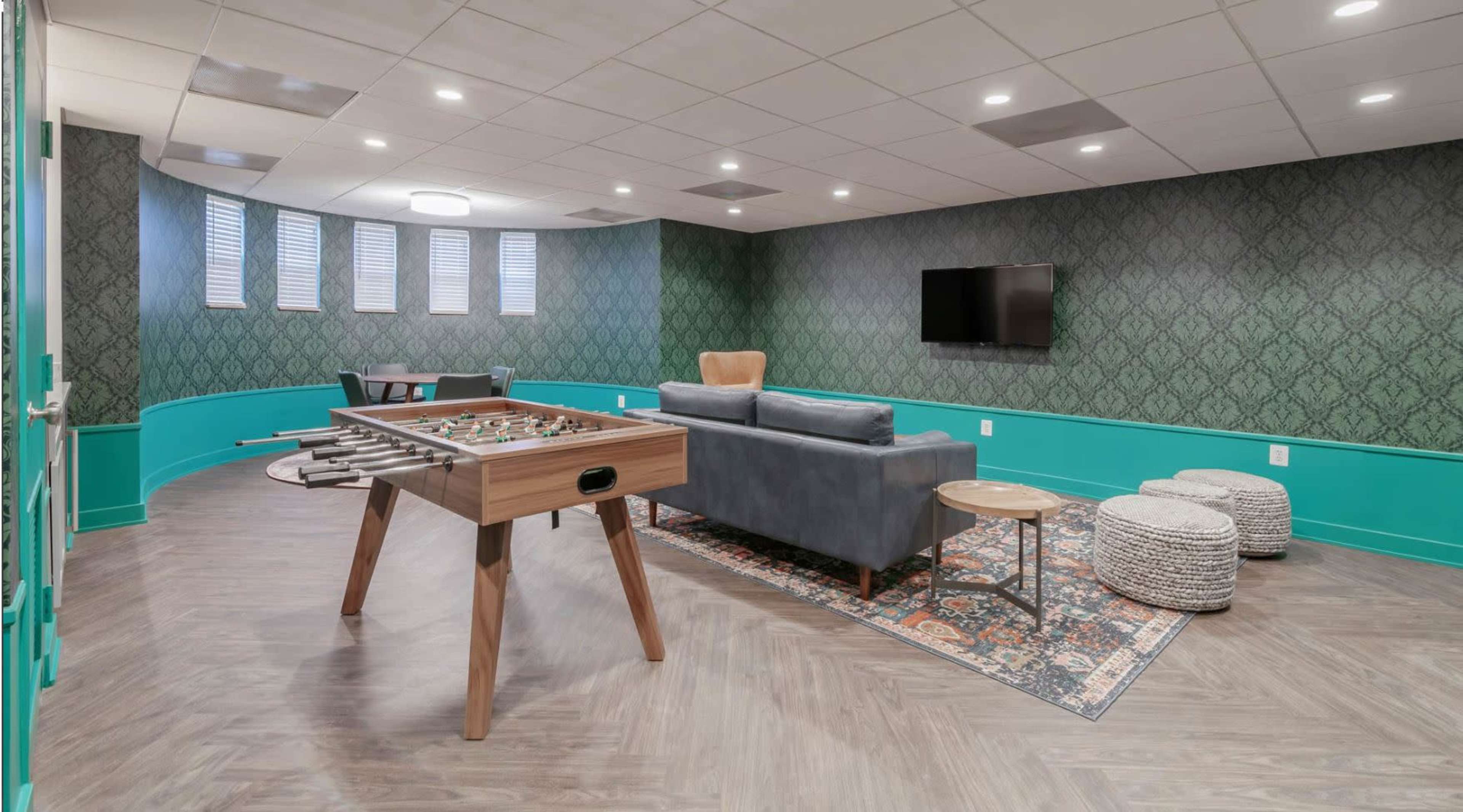 A game room features a foosball table, a gray sofa, and patterned seating against a green wall.