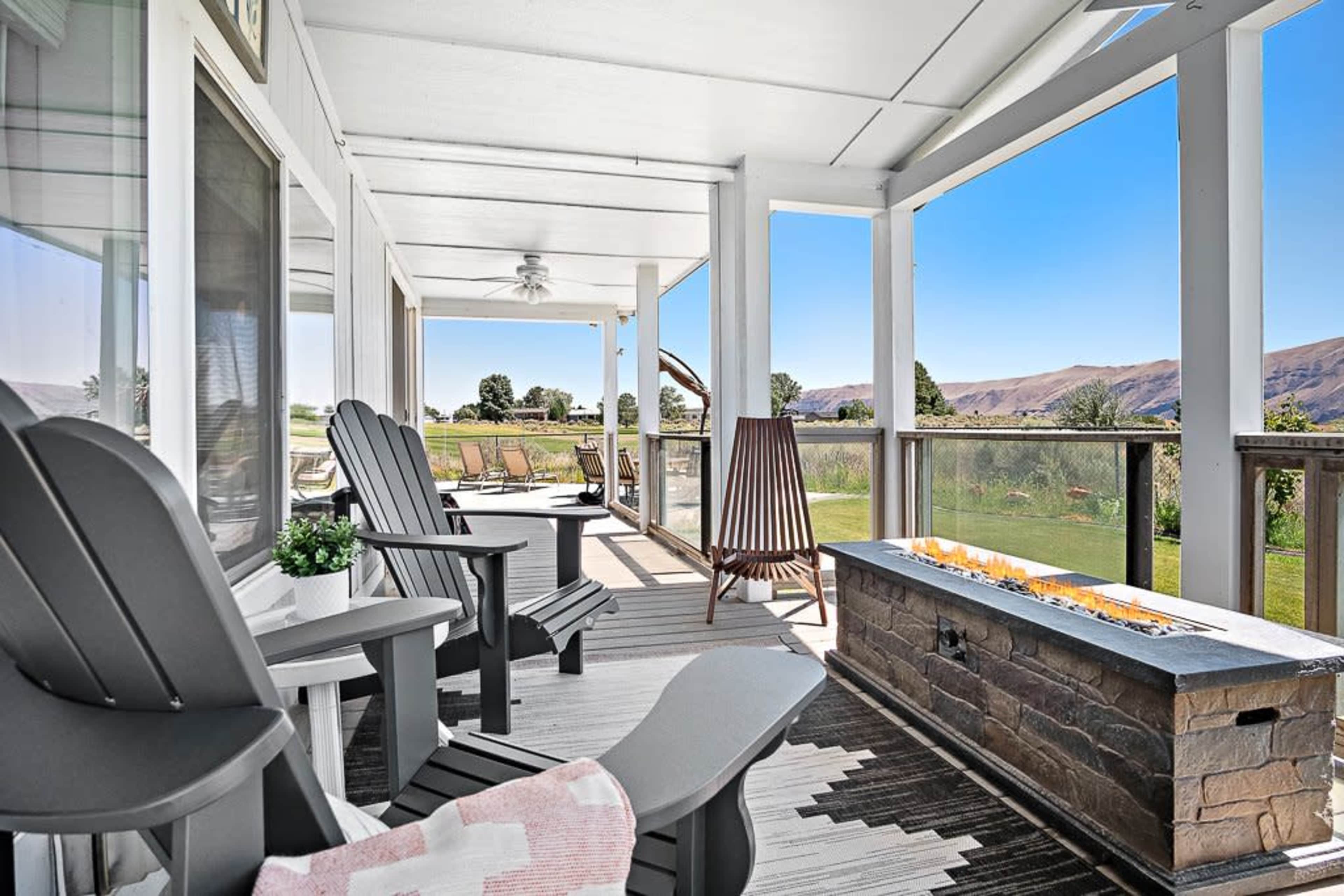 A spacious porch with comfortable chairs, a fire feature, and a view of a landscaped yard and mountains in the background.