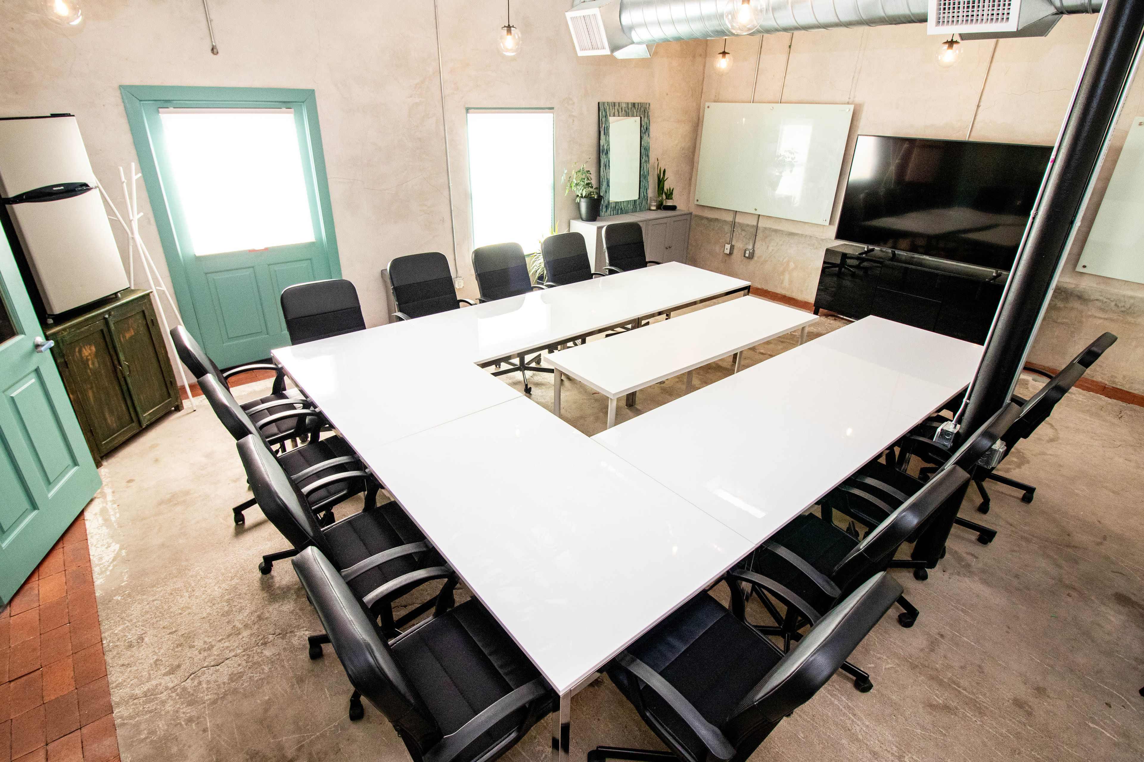 A modern conference room features a large white table arranged in a U-shape with black chairs around it, and includes a television and a kitchenette area.