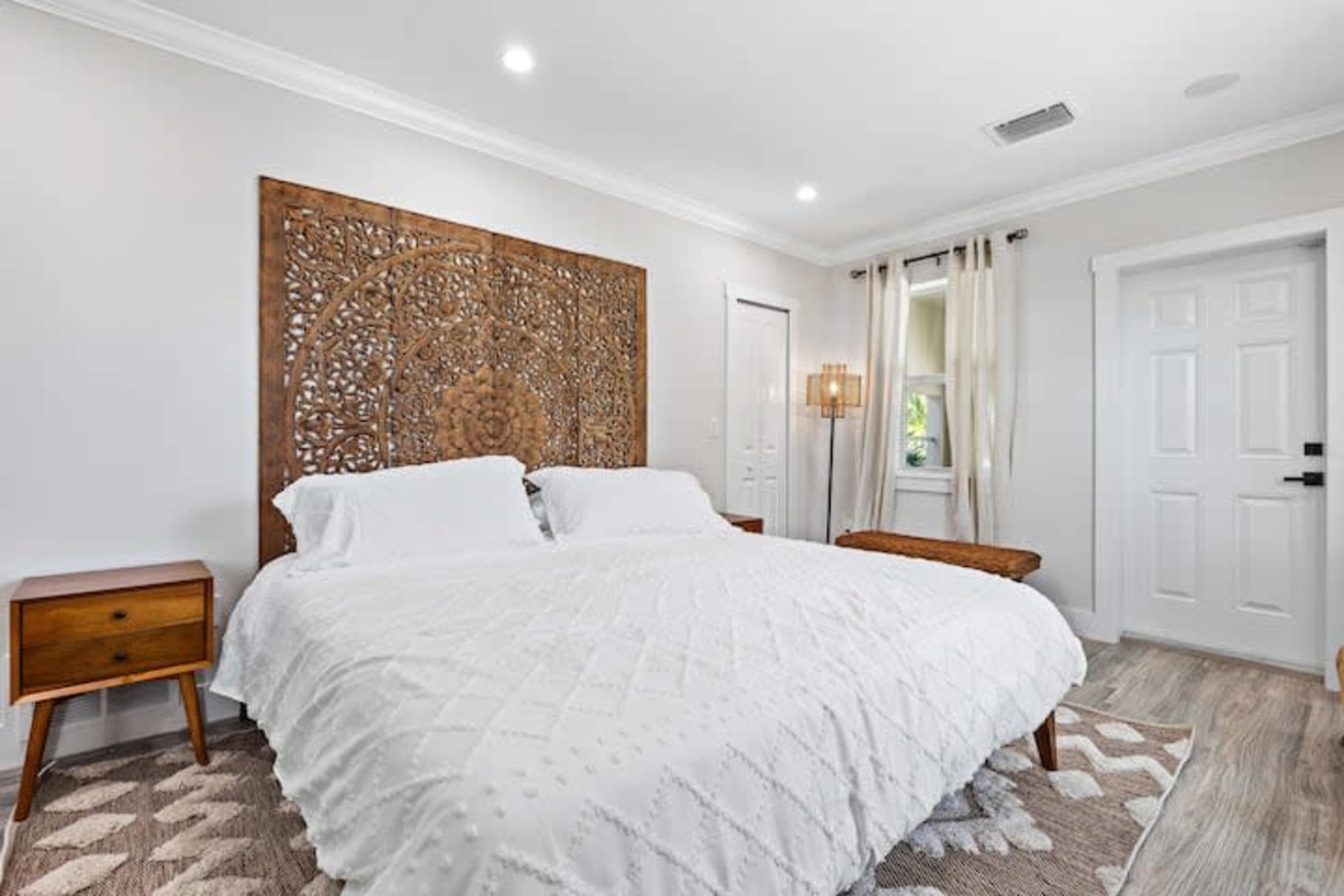 A well-lit bedroom featuring a large carved wooden headboard, a white bedspread, and simple wooden furniture against neutral-colored walls.