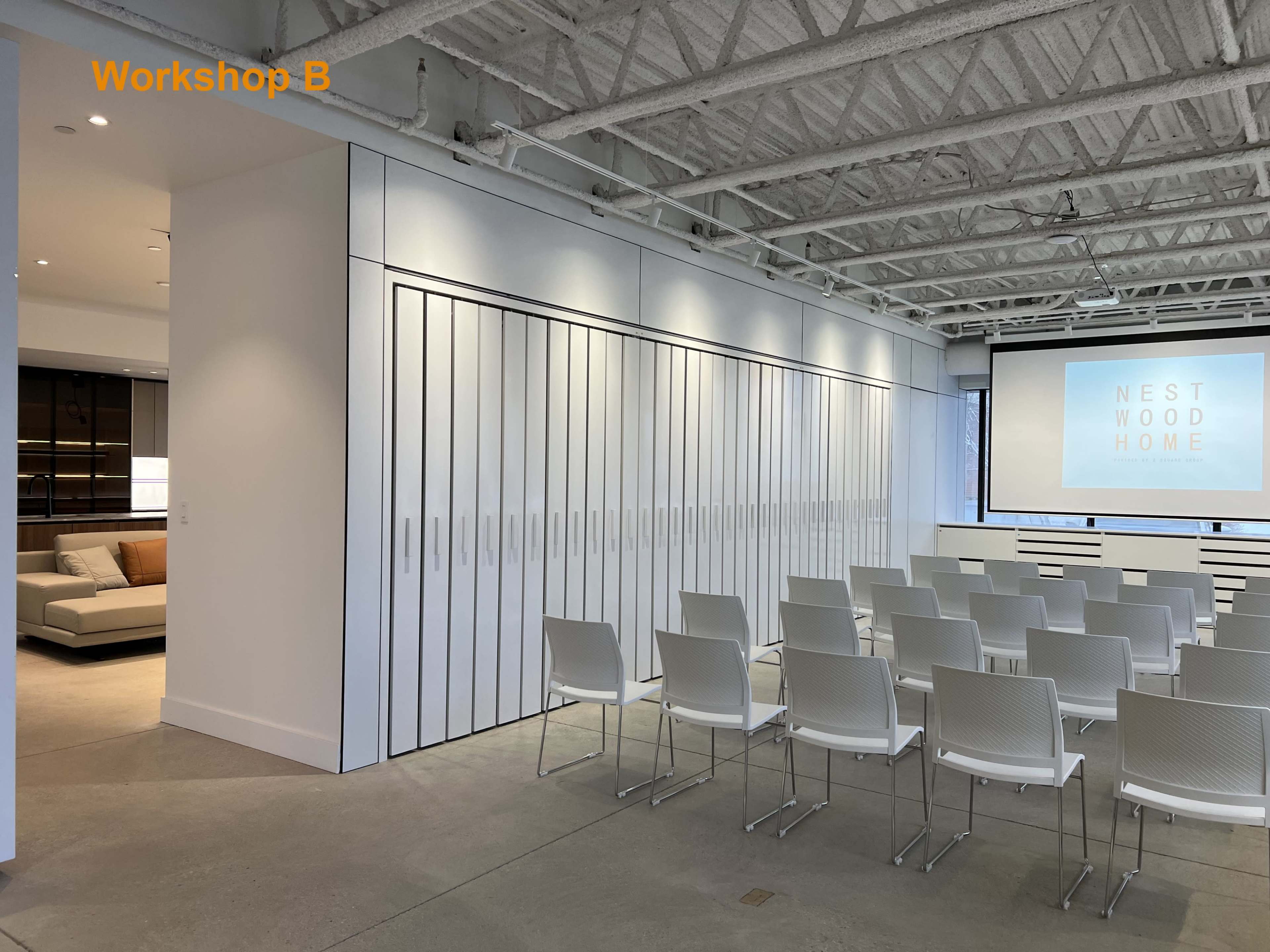 A modern workshop space features rows of white chairs facing a large screen, with white cabinetry lining one wall.
