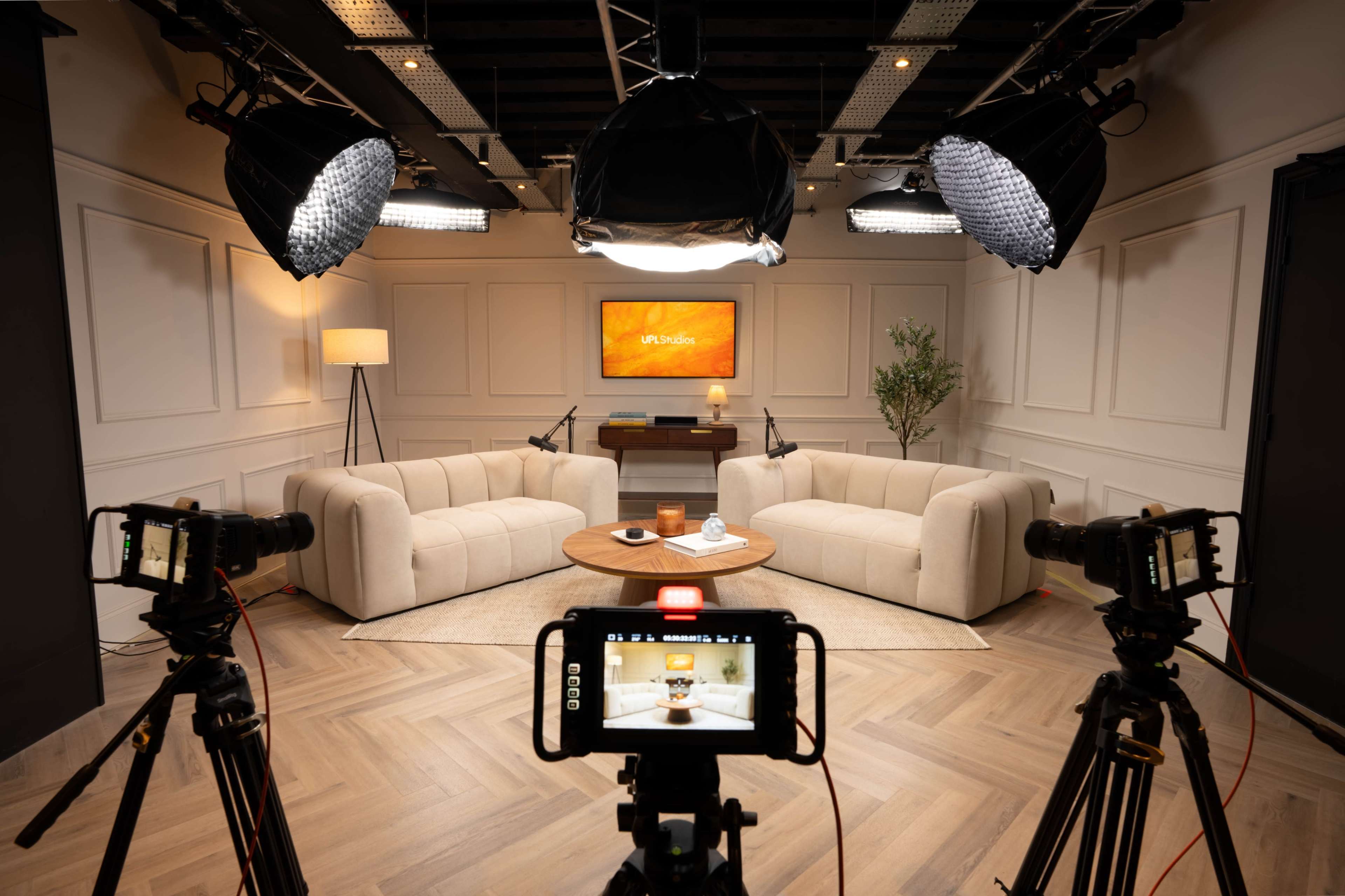 The image shows a well-lit studio setup featuring two beige sofas, a wooden coffee table in the center, and cameras positioned on tripods facing each other.