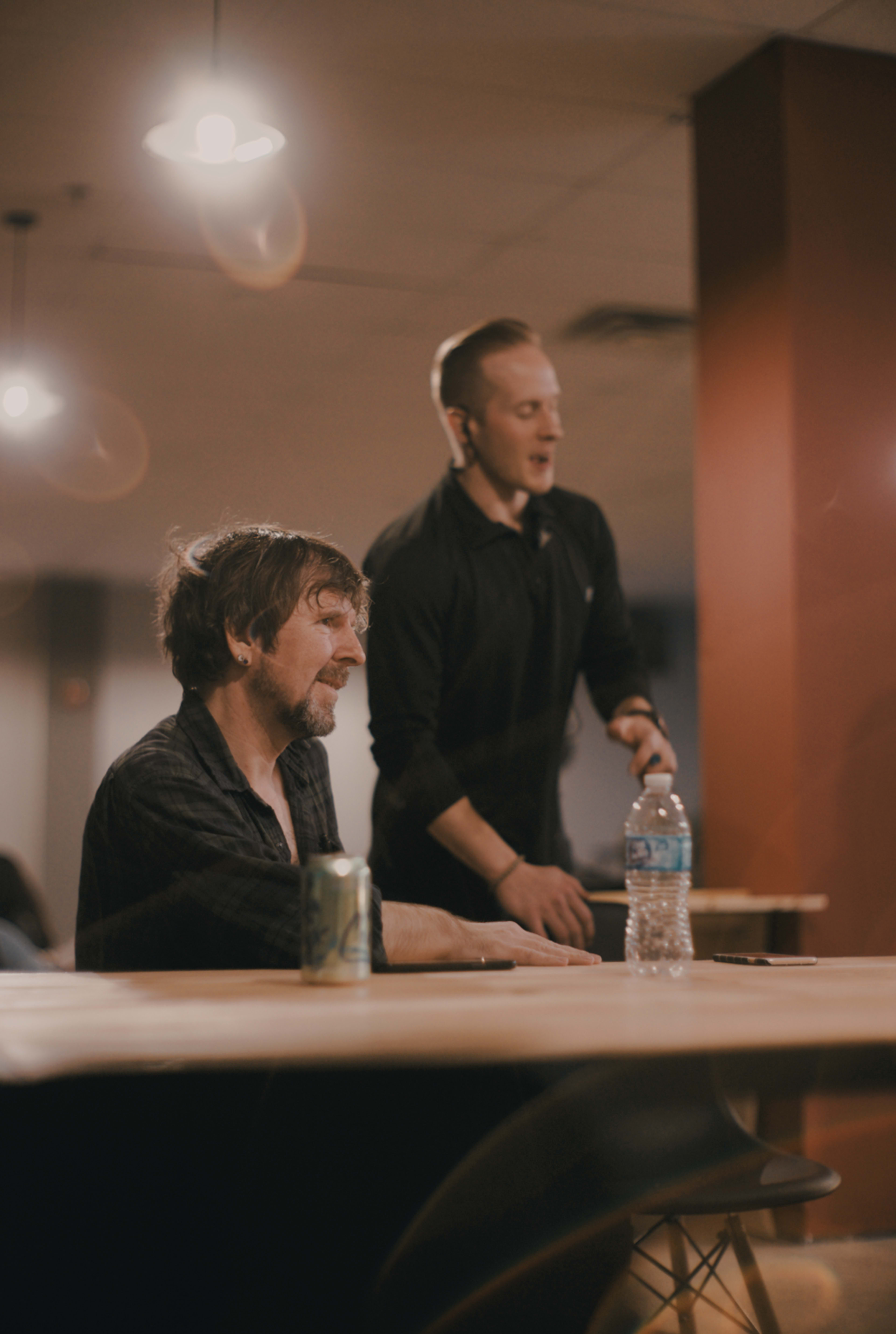 Two men are engaged in conversation at a table, with one standing and the other seated, in a dimly lit room.