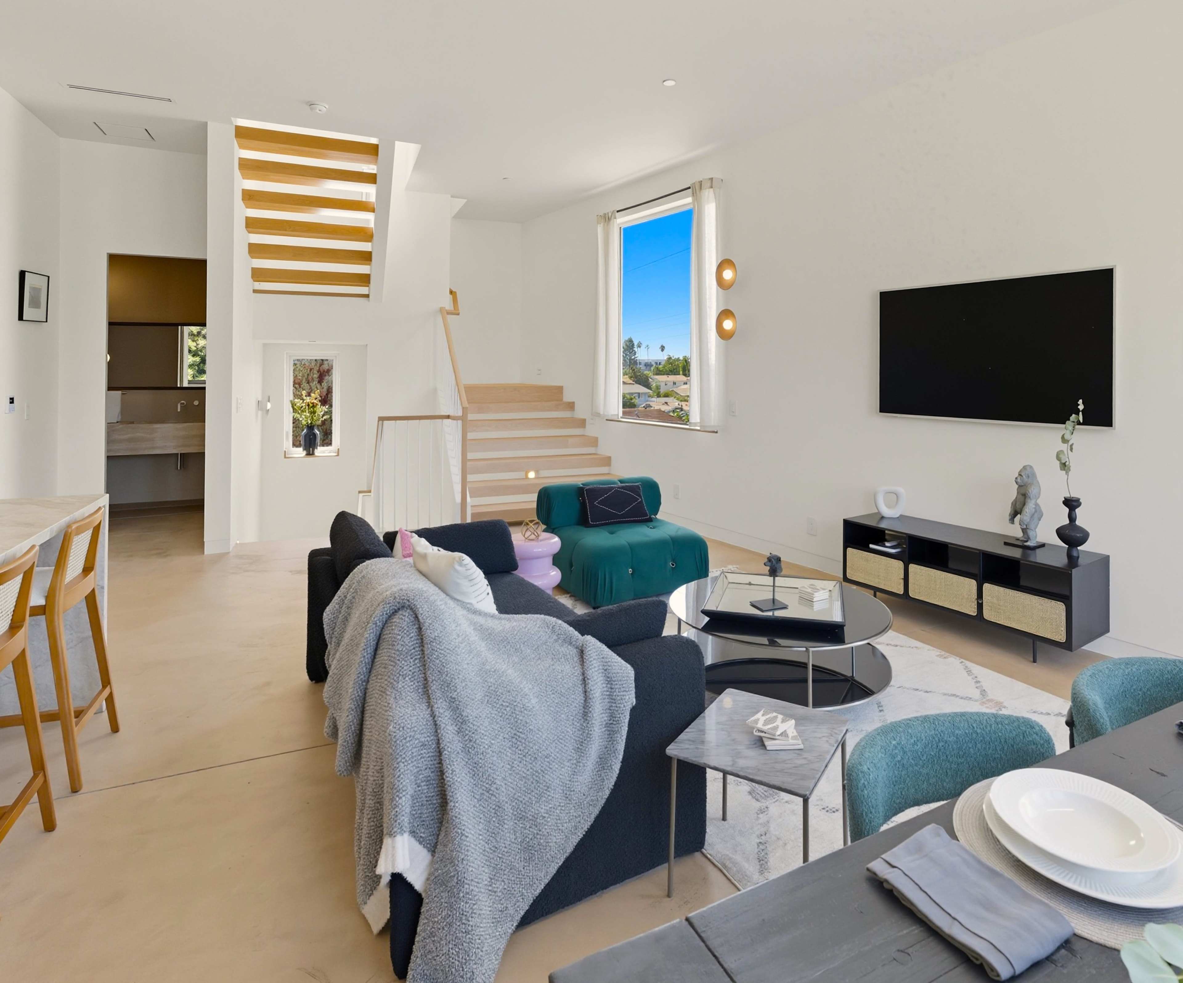 The image shows a modern, open-concept living space featuring a living room with a couch, a coffee table, and dining area, along with a staircase leading to an upper floor.