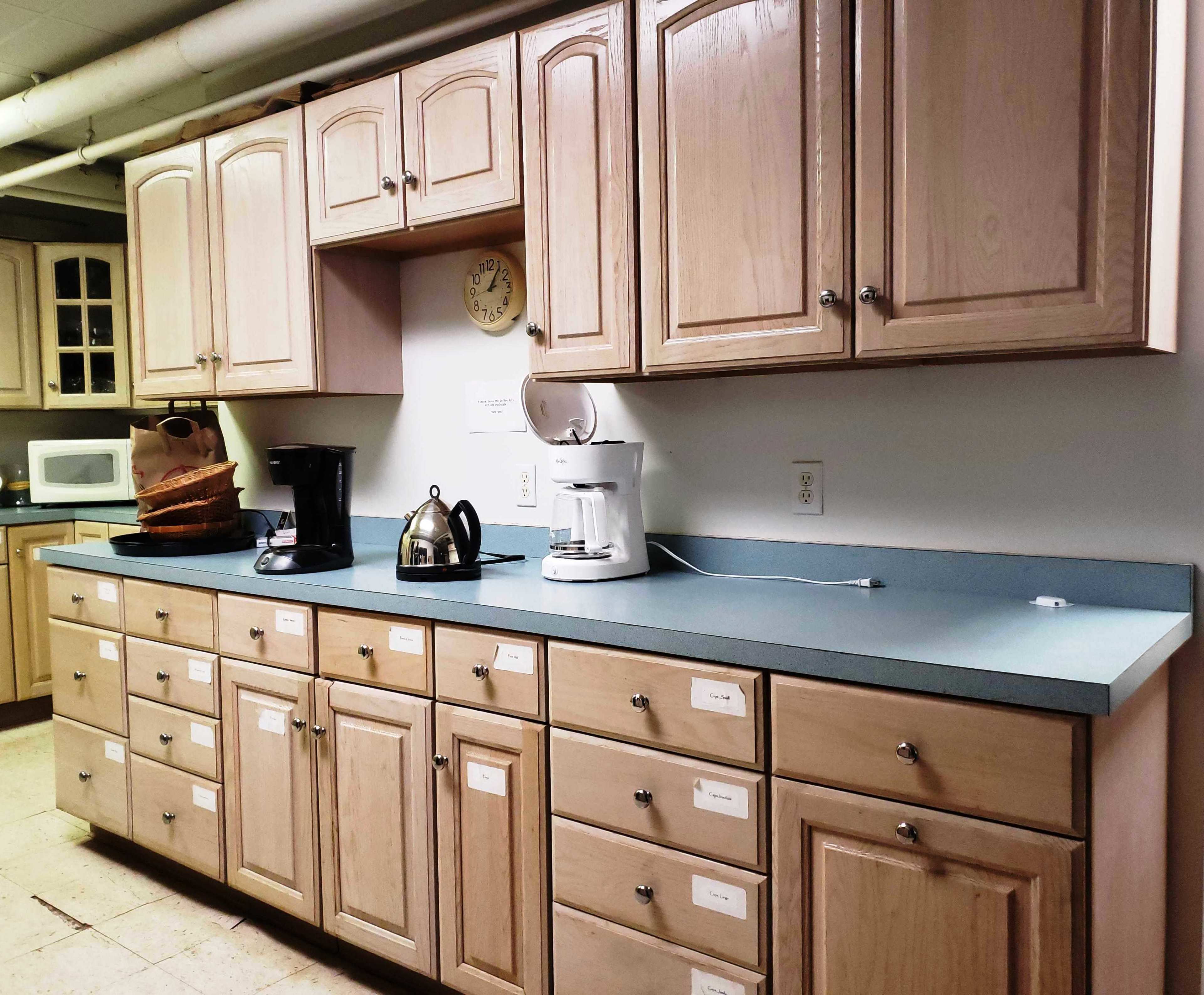 A kitchen counter is lined with cabinets and appliances, including a coffee maker, kettle, microwave, and a clock.