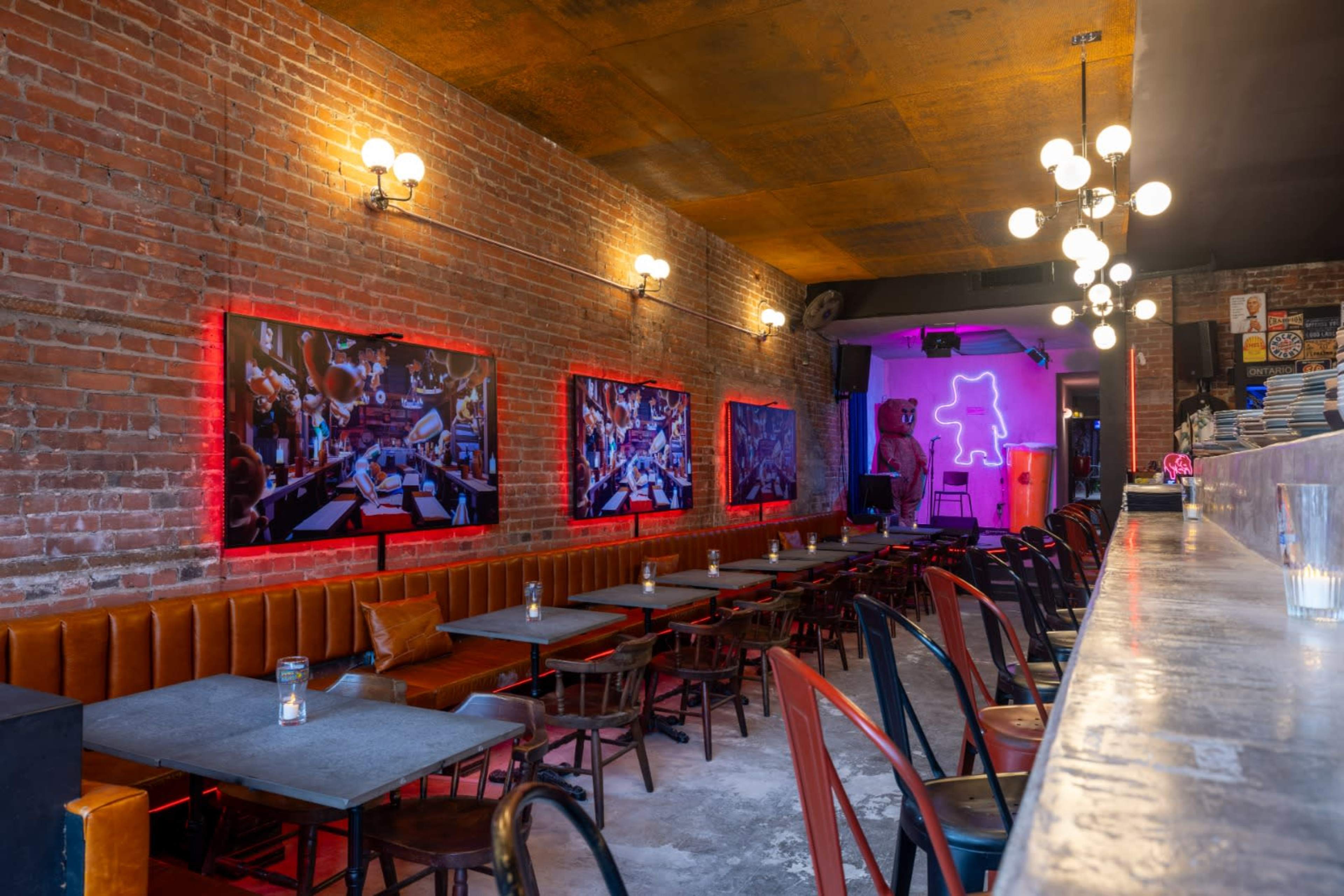 The image shows a restaurant interior with exposed brick walls, a long bar area, and rows of tables with chairs, illuminated by colorful lights and artwork on the walls.