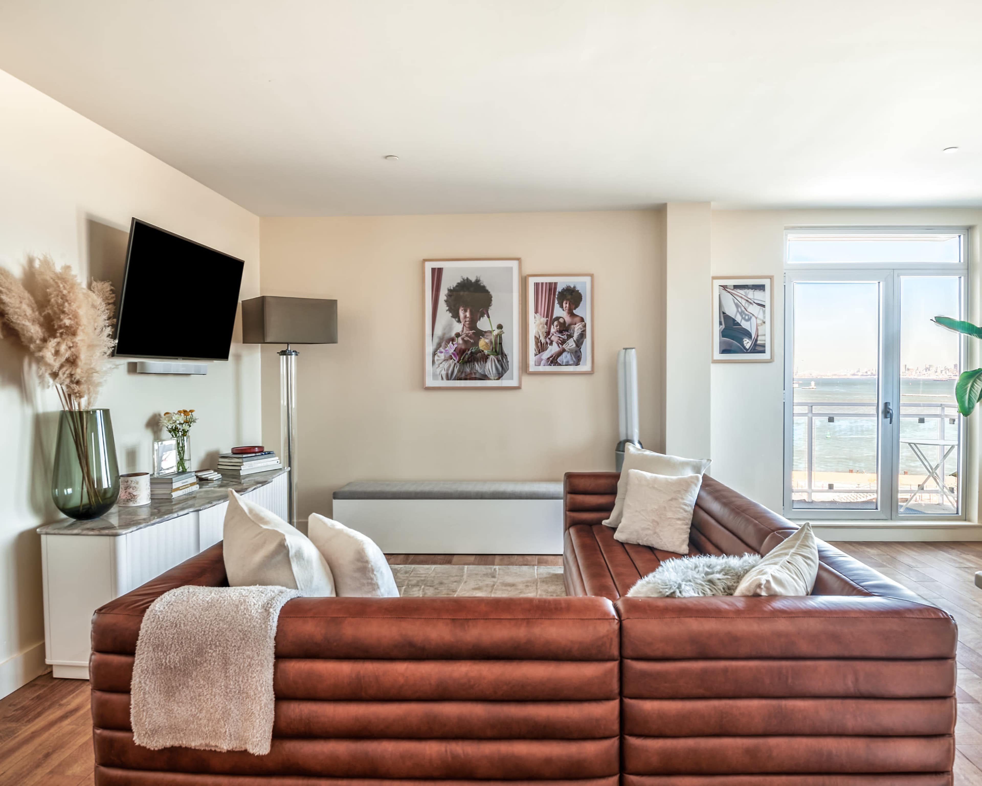 A modern living room featuring a brown sectional sofa, a flat-screen TV mounted on the wall, and large windows with a water view.
