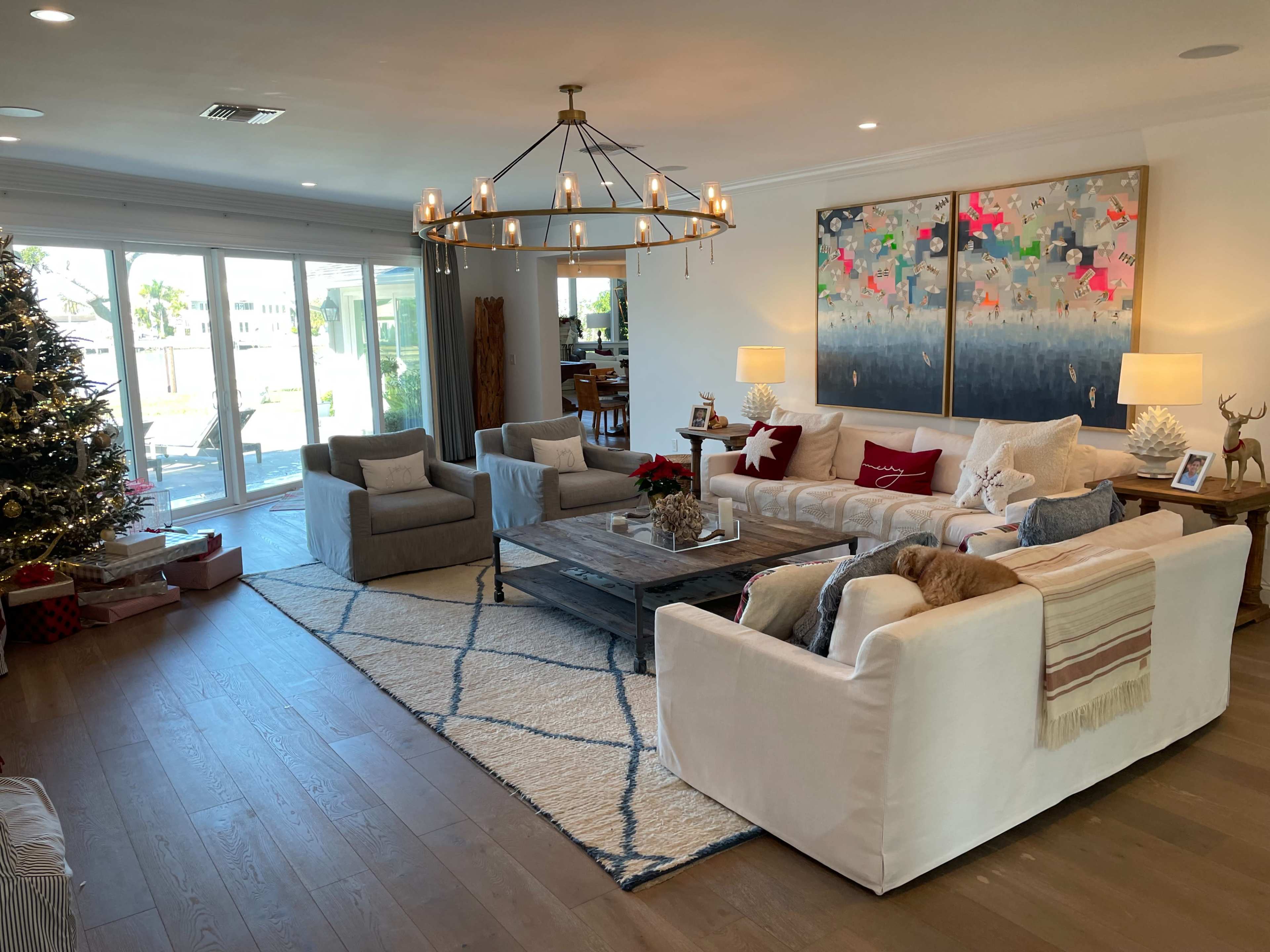 A modern living room decorated for the holidays, featuring a Christmas tree, two sofas, and a large coffee table, with ample natural light coming from sliding glass doors.