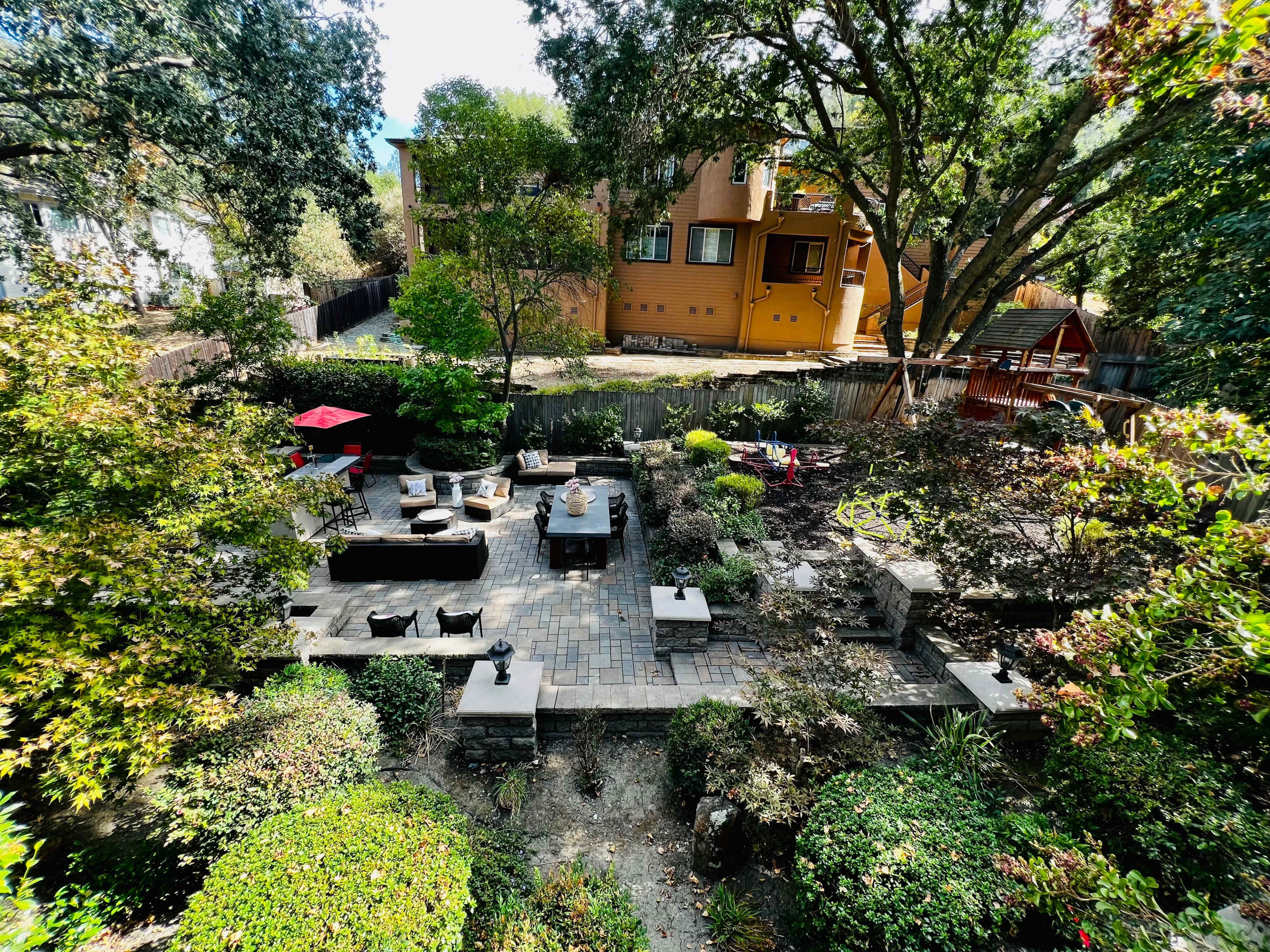 A landscaped garden with stone pathways, seating areas, and various plants, set against a backdrop of a two-story house and trees.