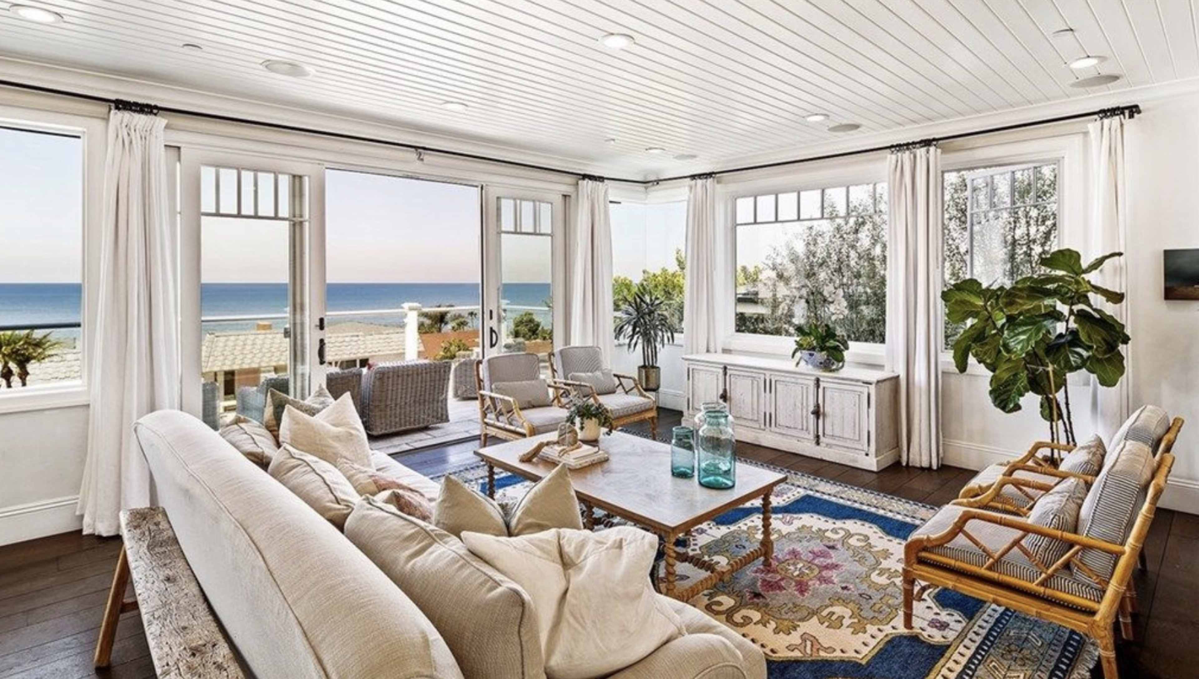A modern living room with large windows overlooking the ocean, furnished with a light sofa, wooden chairs, and a large area rug.