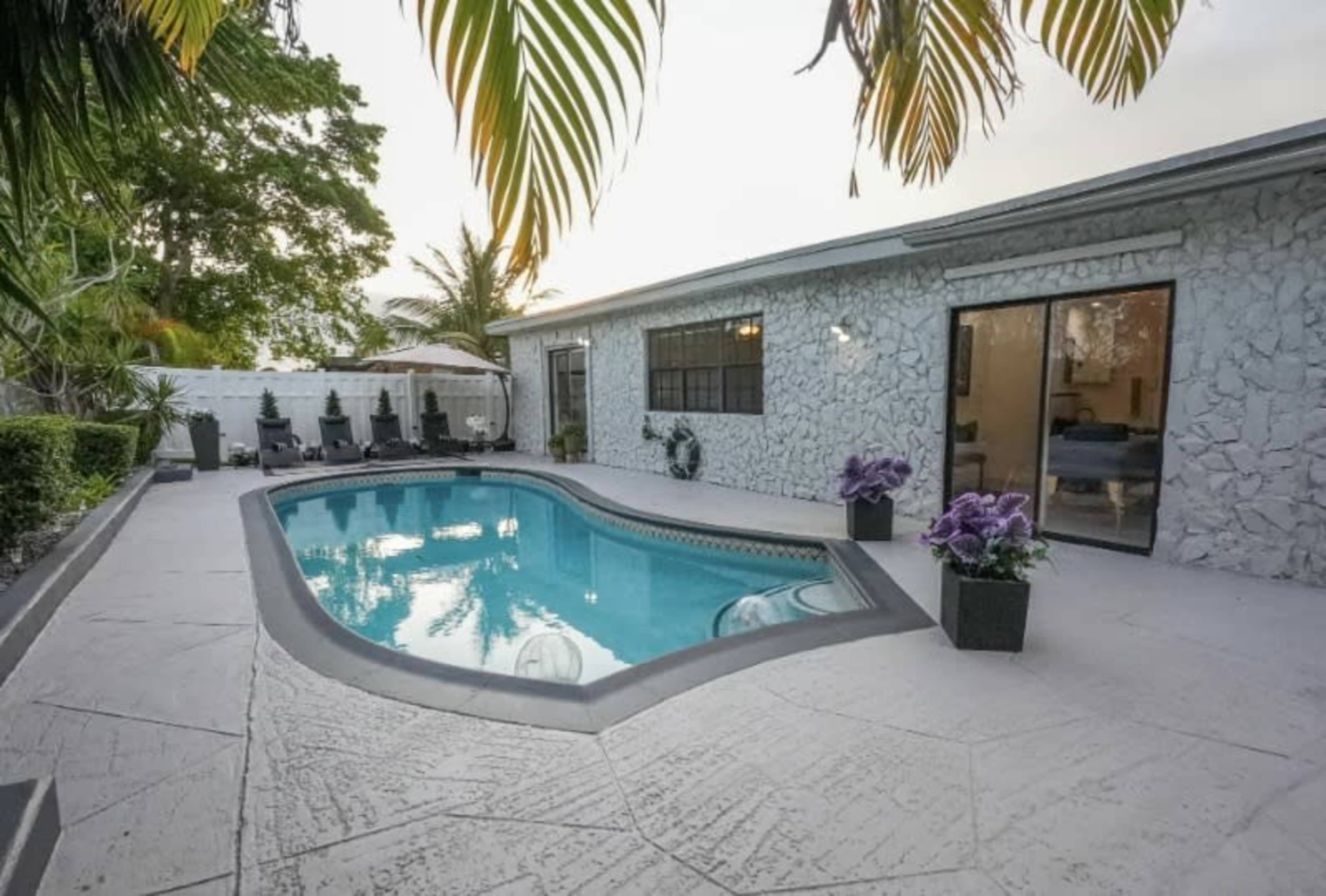 The image shows a backyard pool area surrounded by tropical plants and a textured stone exterior of a house.