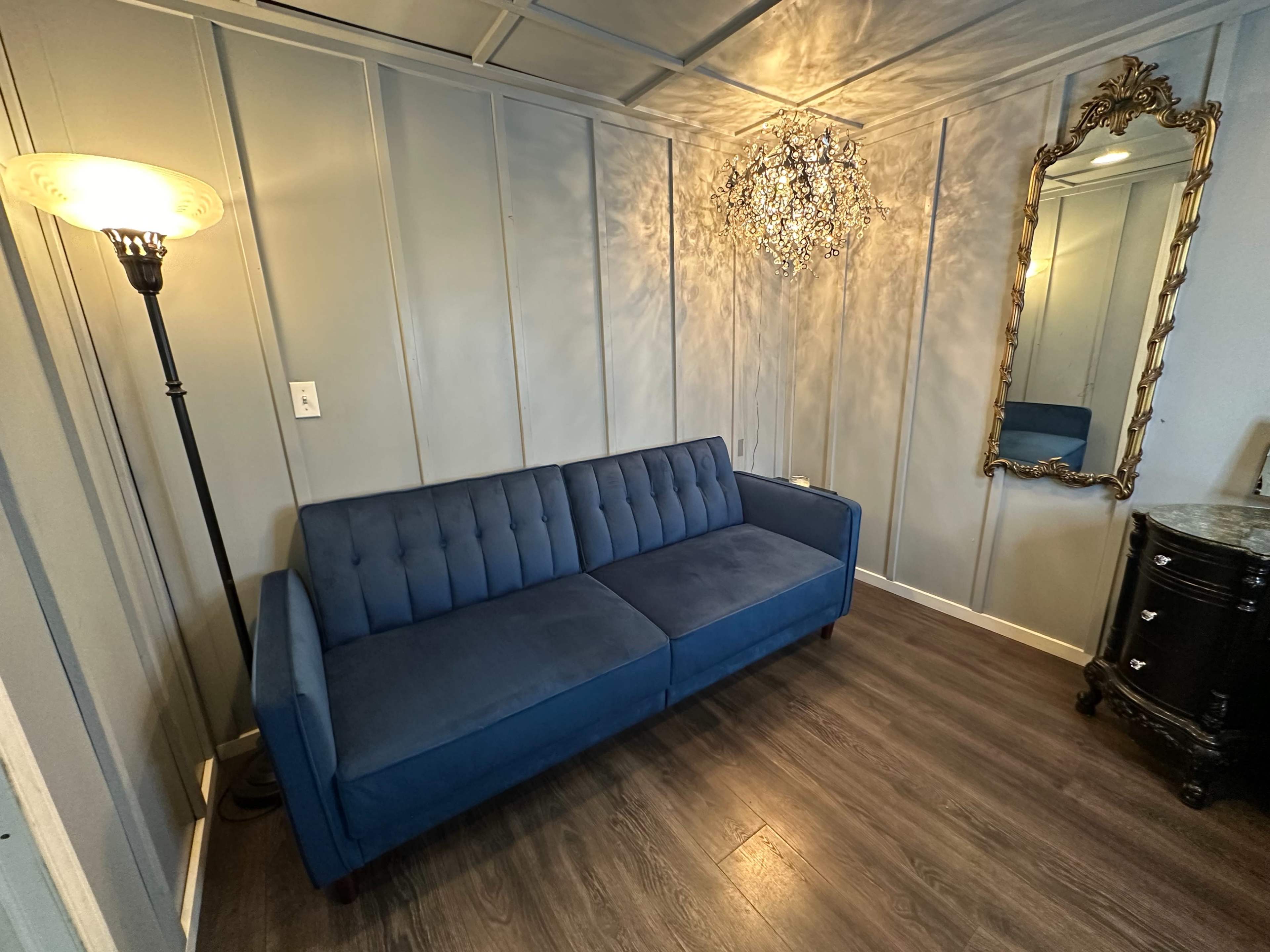 A blue tufted sofa in a room with gray paneling, a decorative chandelier, and a vintage mirror, alongside a floor lamp and a small black cabinet.