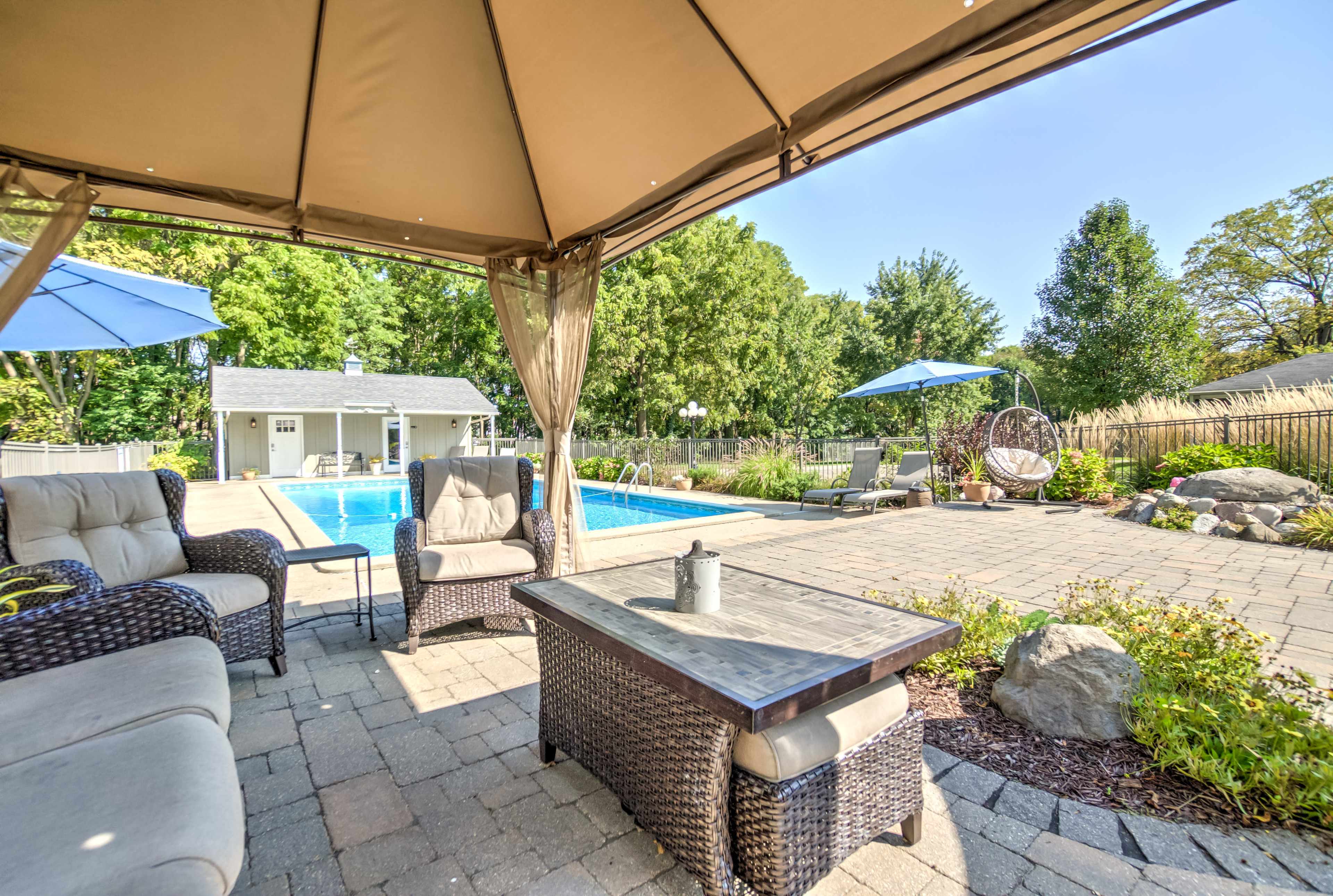 A shaded seating area with wicker furniture overlooks a swimming pool surrounded by greenery and lounge chairs.
