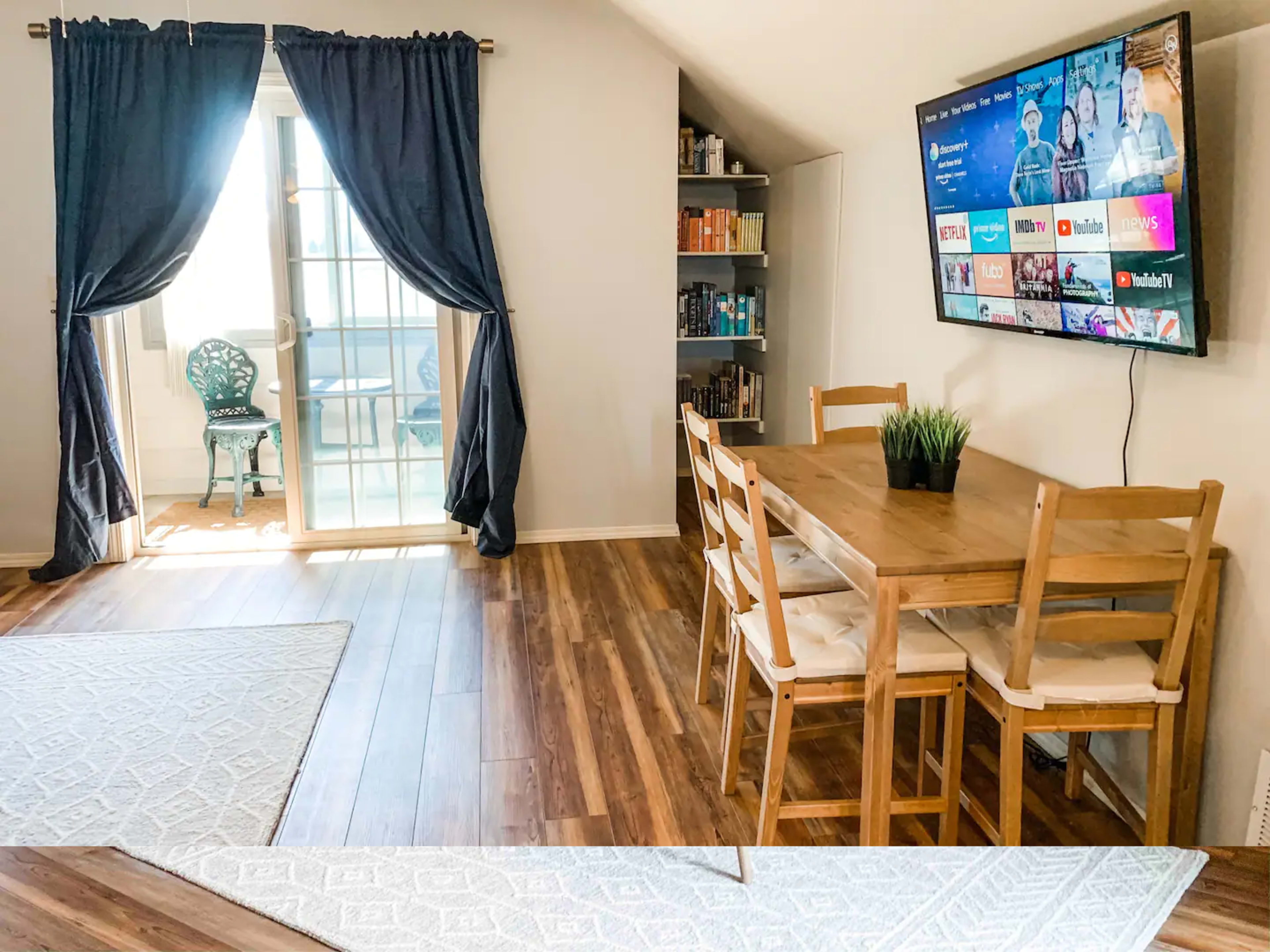 A cozy dining area with a wooden table and chairs, a bookshelf filled with books, a wall-mounted TV, and large curtains framing a patio door that opens to a small balcony.
