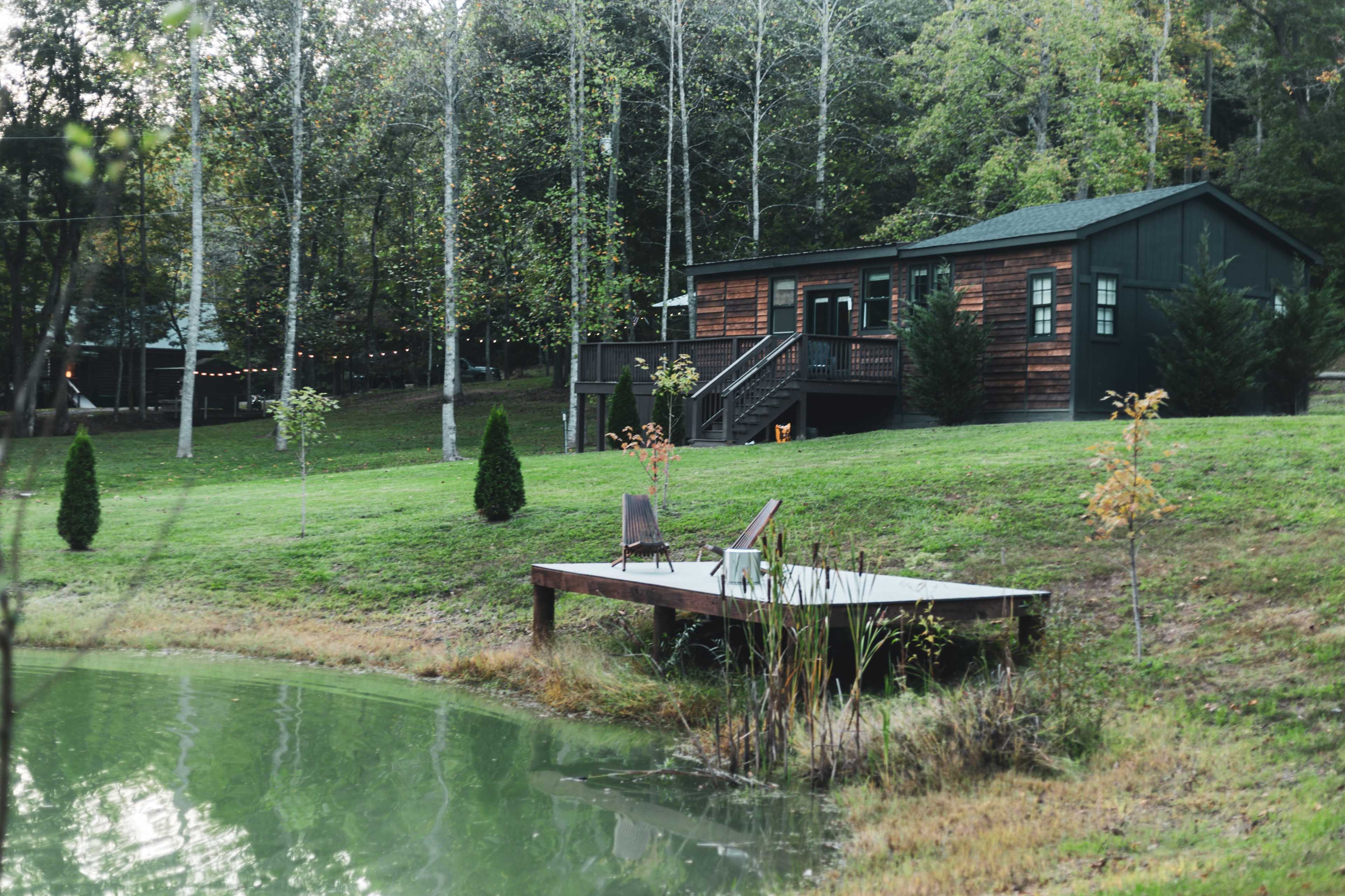 A wooden cabin sits on a grassy hill beside a green pond, with a small dock extending into the water.
