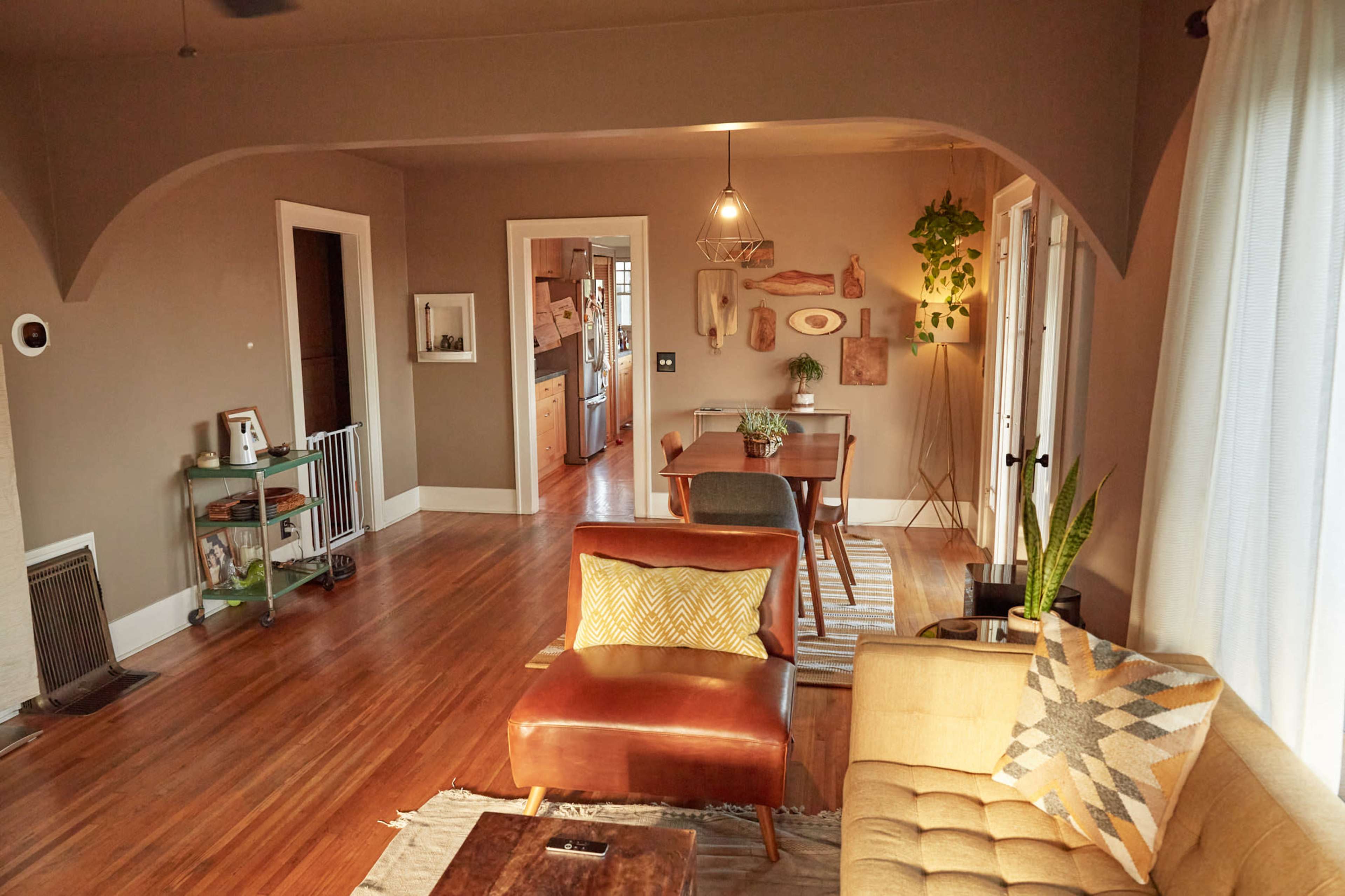 A living room with wooden flooring, a yellow sofa, a brown armchair, a dining table in the background, and plants placed around the space.