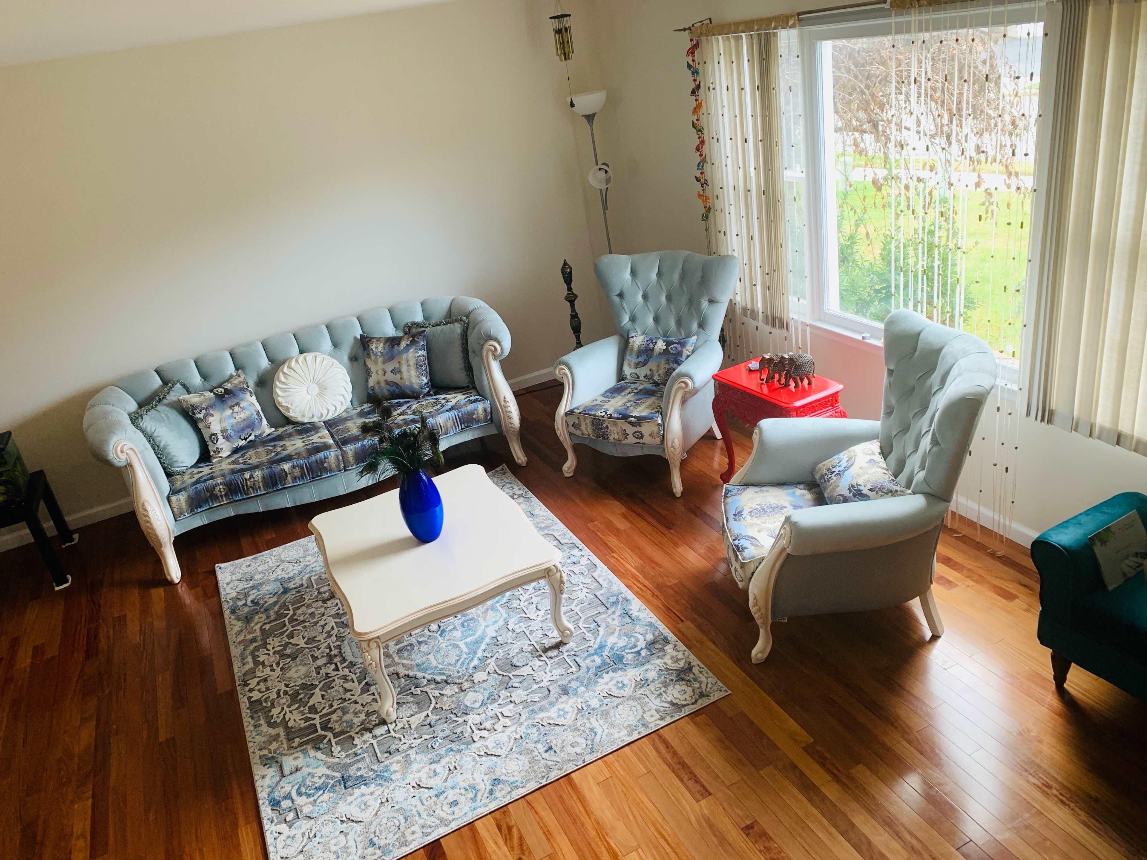 The image shows a living room with a light-colored couch, two matching armchairs, a white coffee table, and a decorative rug on hardwood flooring near a window with white curtains.