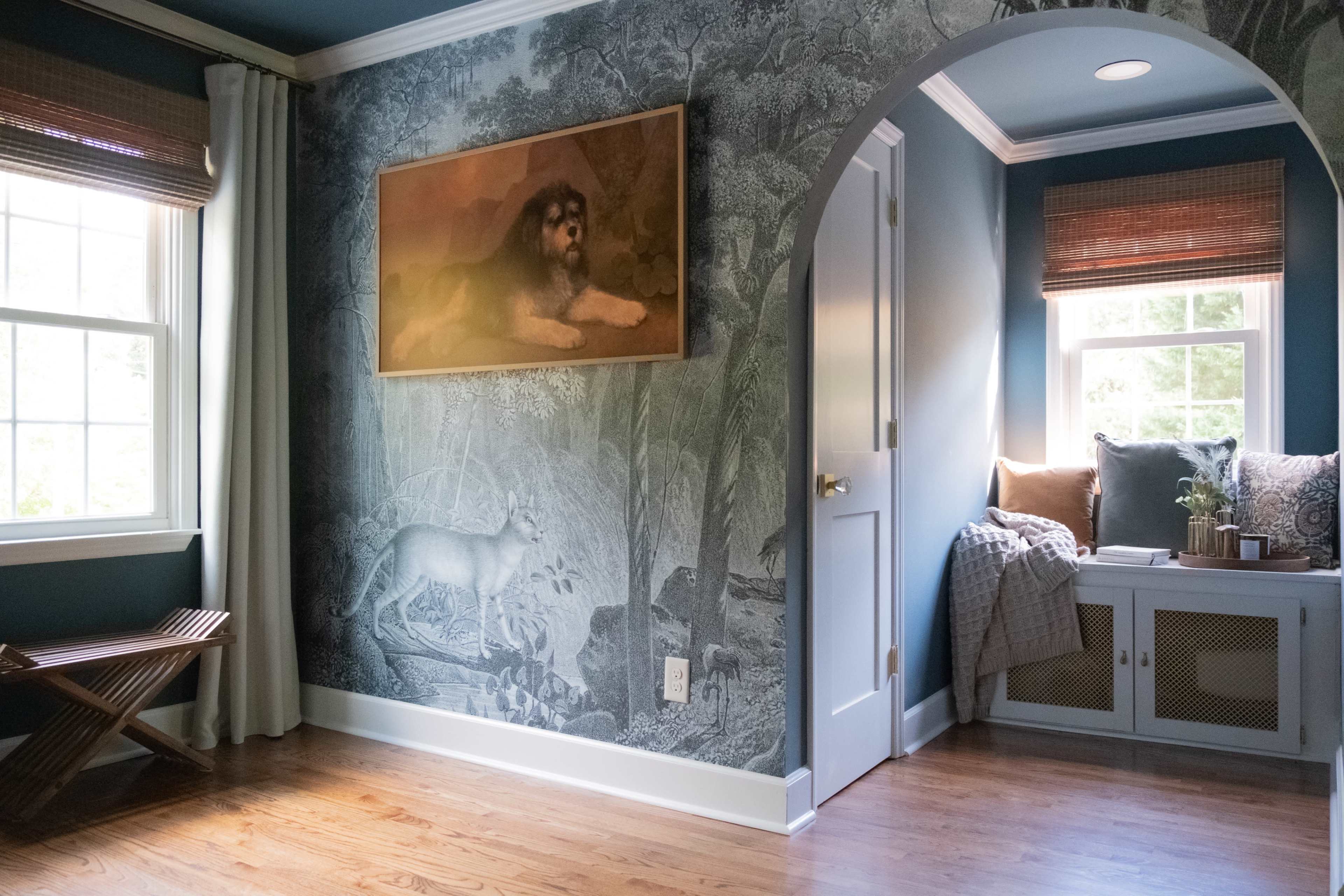 A room with dark blue walls decorated with a large mural, featuring a large dog and a white deer, and a cozy nook with cushions and plants next to a window.