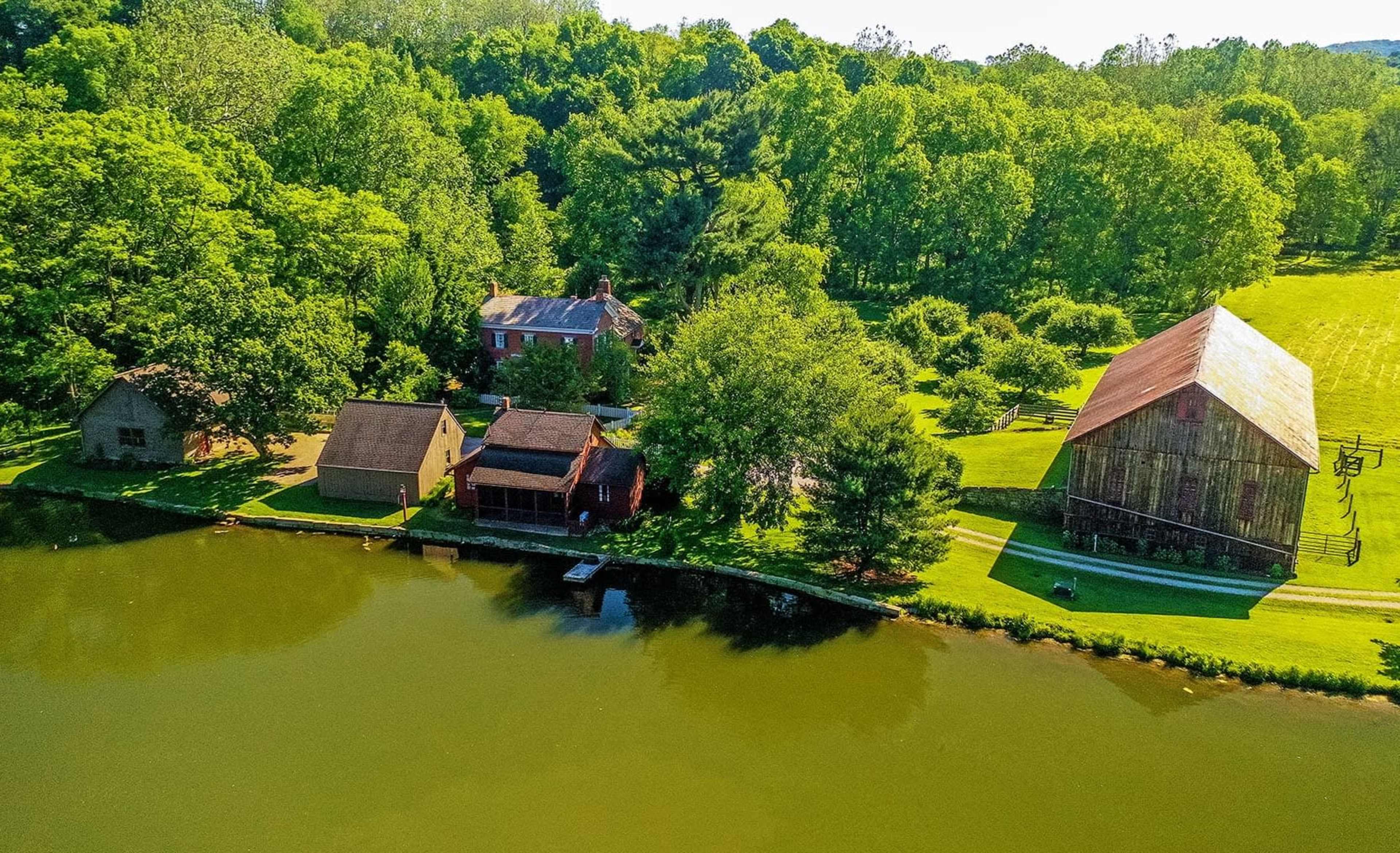 A rural landscape featuring several buildings, including a house and a barn, along the edge of a calm pond surrounded by green trees.