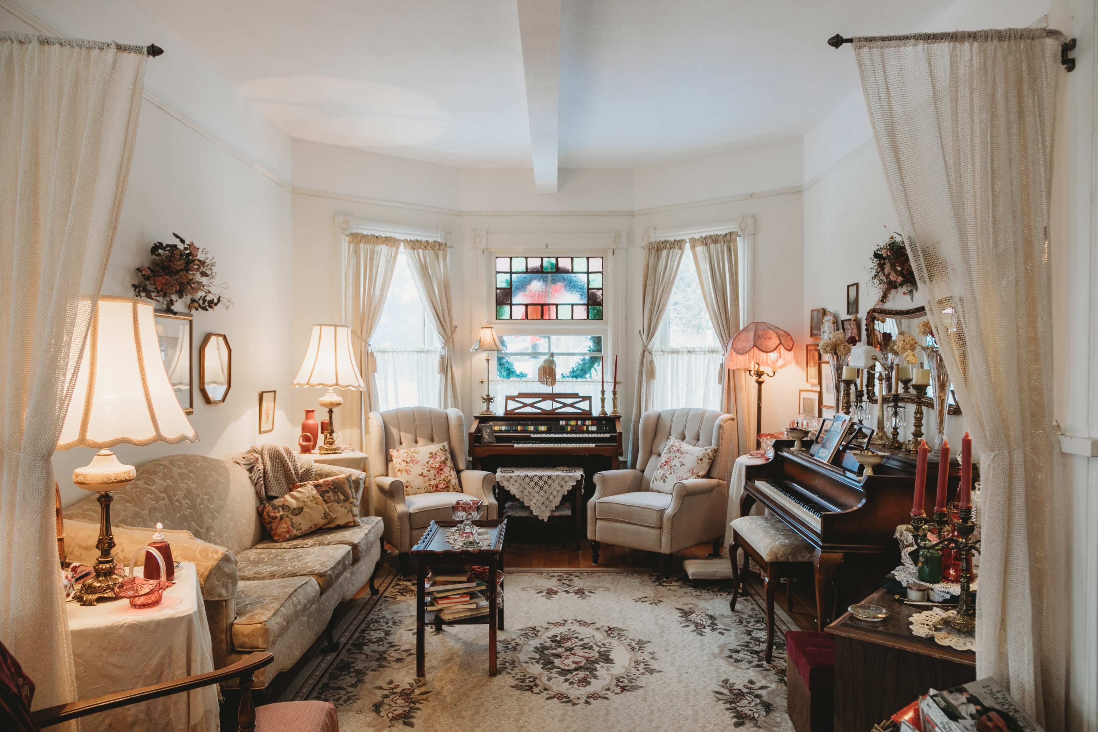 The image shows a cozy living room with two armchairs, a sofa, a piano, and large windows adorned with sheer curtains, illuminated by lamps.