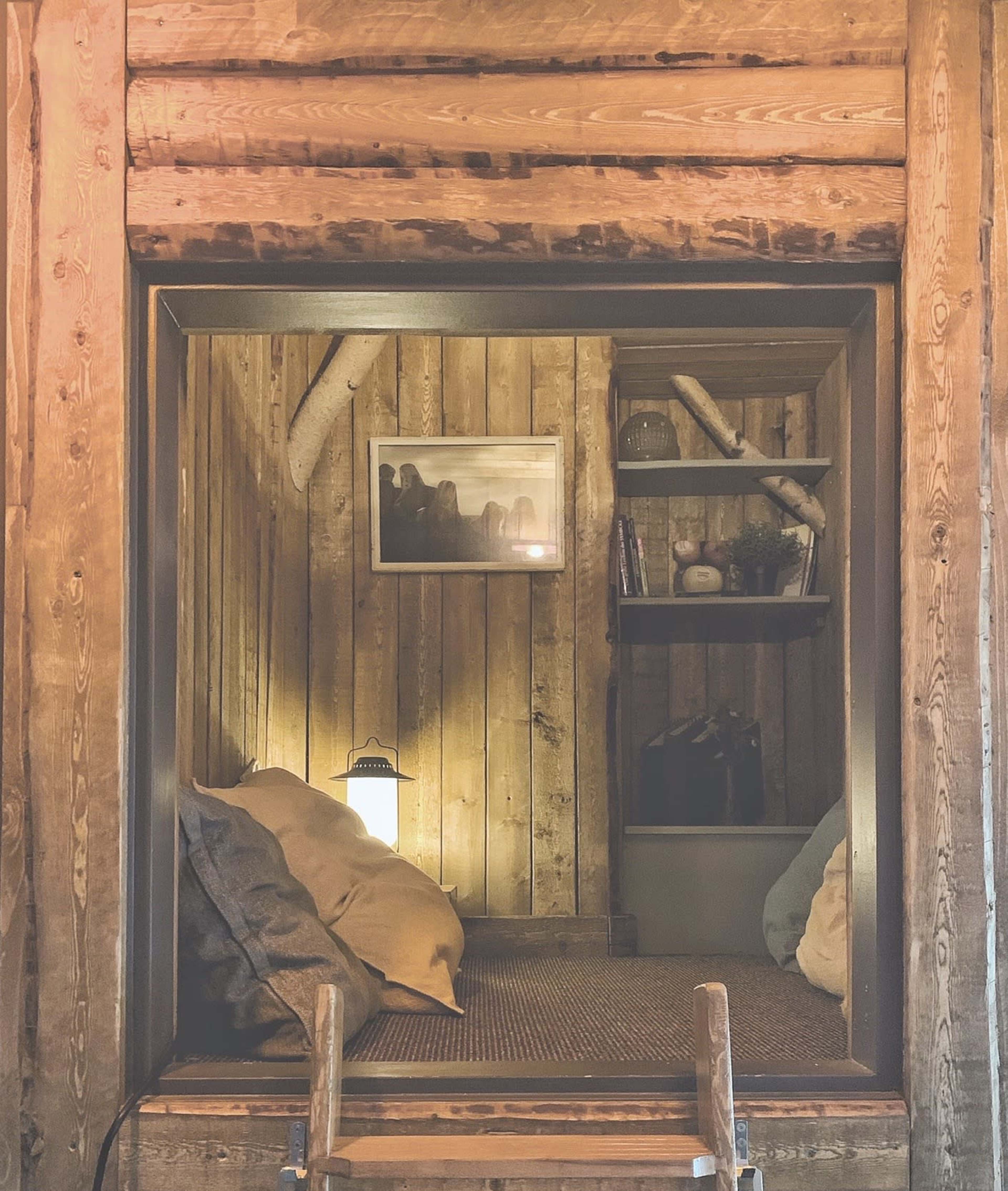 A cozy nook with wooden walls contains a bed, a lamp, and a shelf with books.
