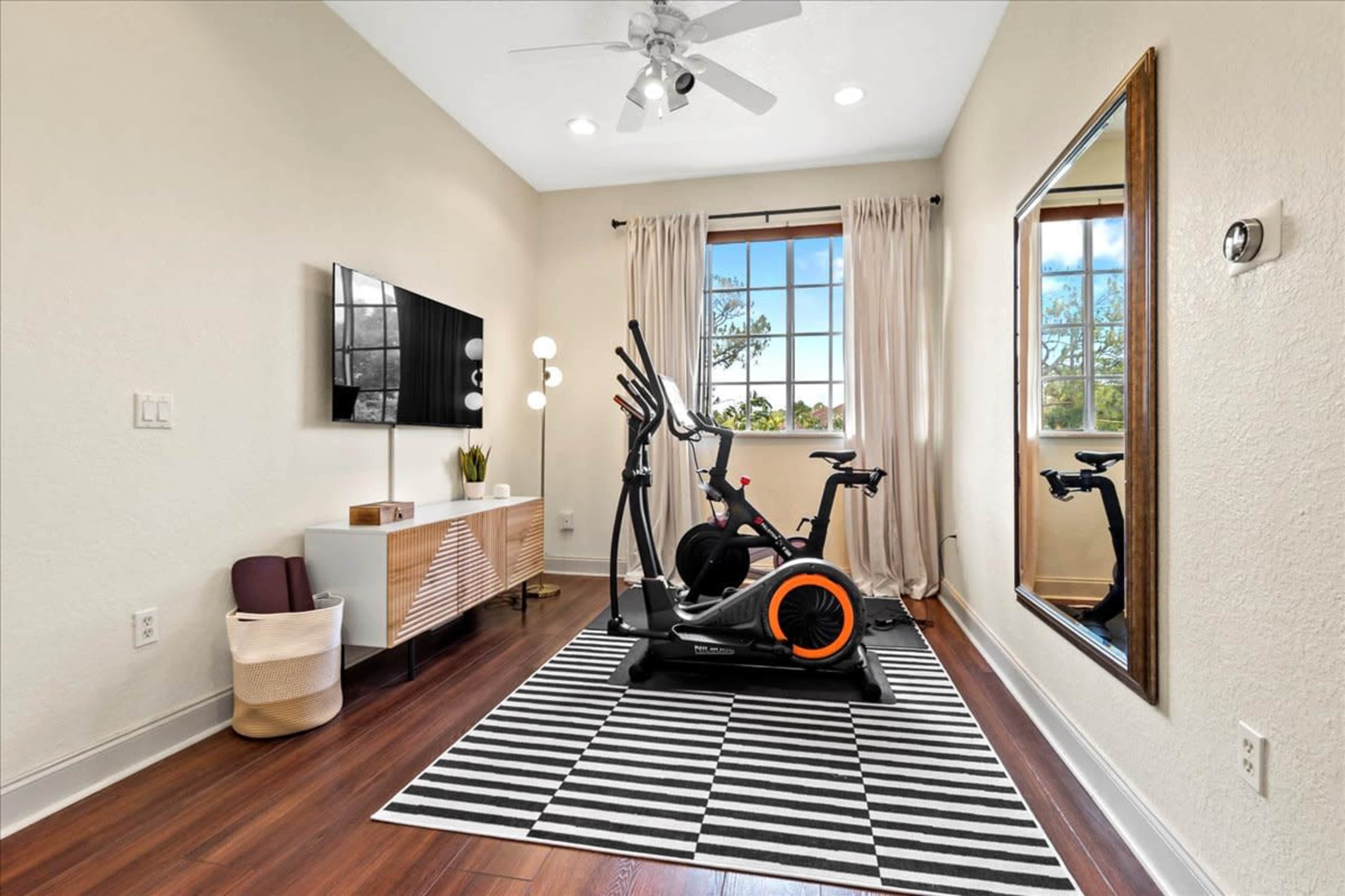 A home gym featuring an elliptical machine, a large mirror, a TV mounted on the wall, and a striped rug on a wooden floor.