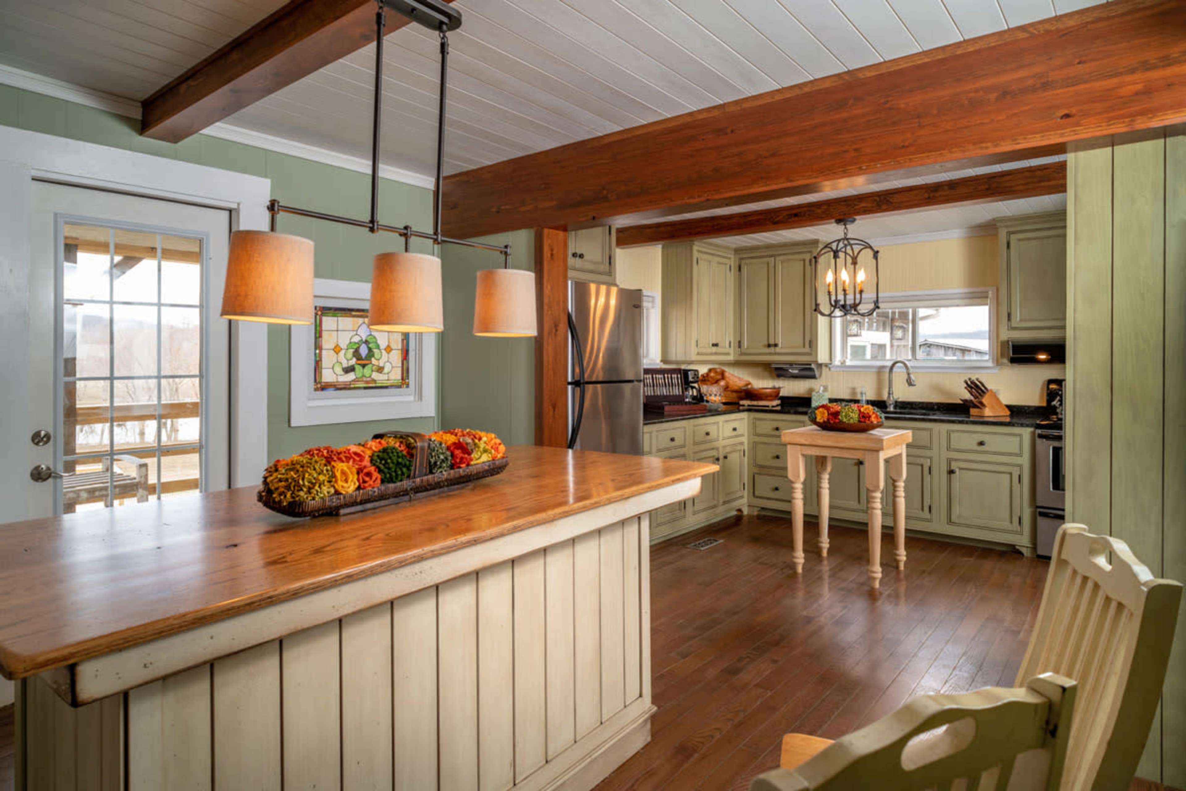 The kitchen features a central island with a wooden countertop, surrounded by light-colored cabinetry, pendant lighting, and a view of an outdoor deck through a glass door.