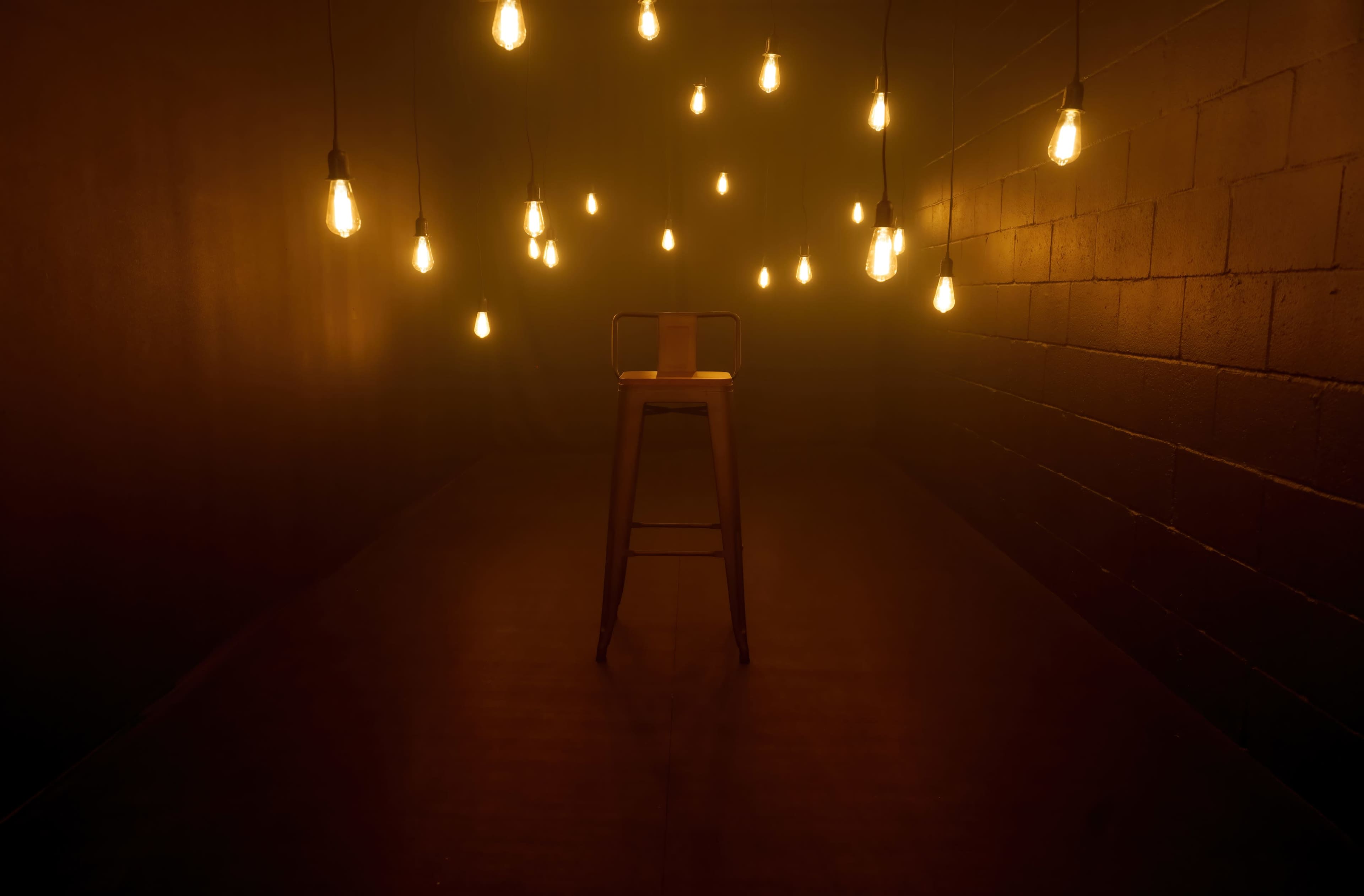 A single wooden stool is positioned in a dimly lit room surrounded by hanging light bulbs.