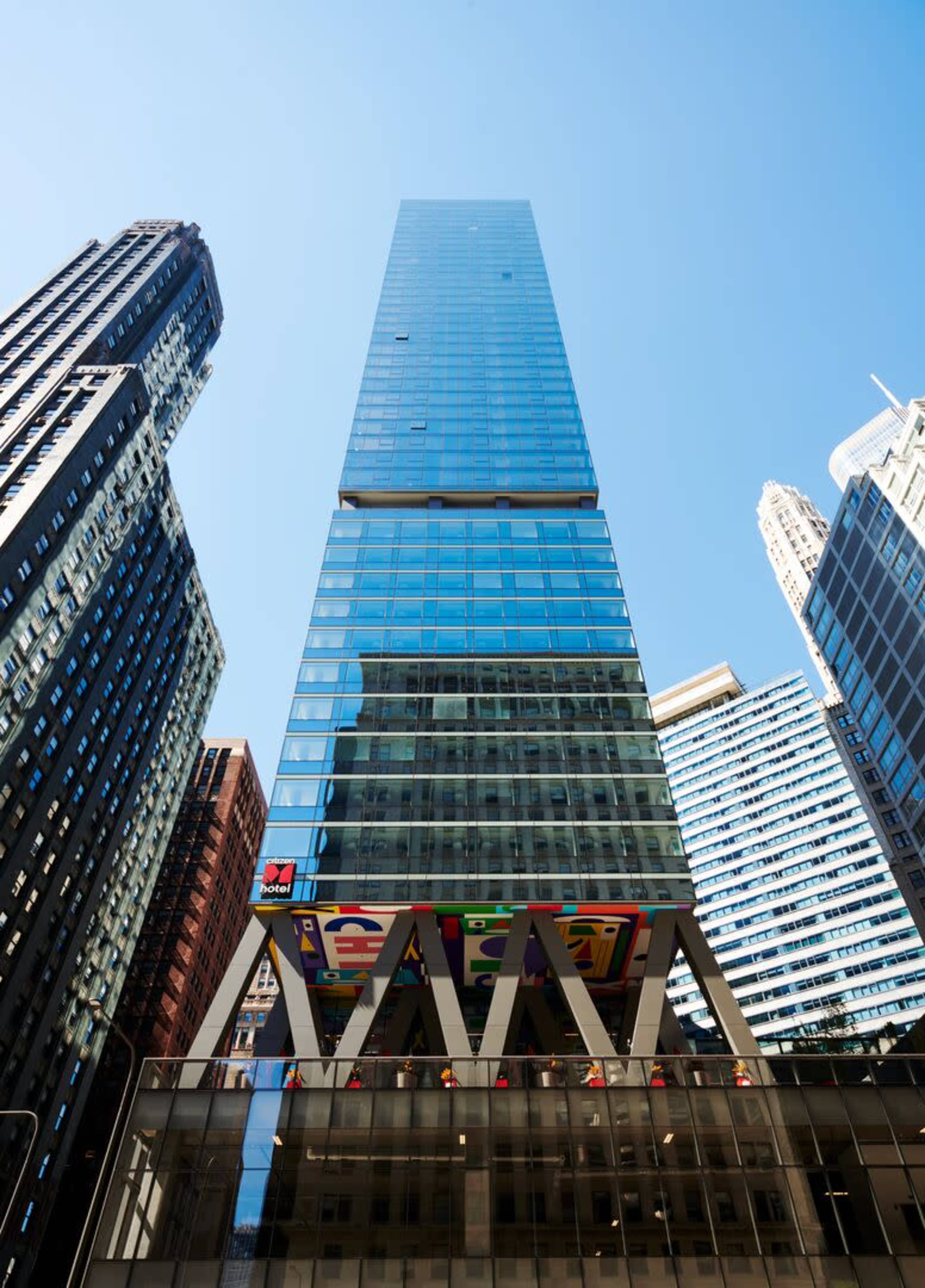 A tall glass skyscraper stands between shorter buildings, with a colorful awning supported by large beams at its base.
