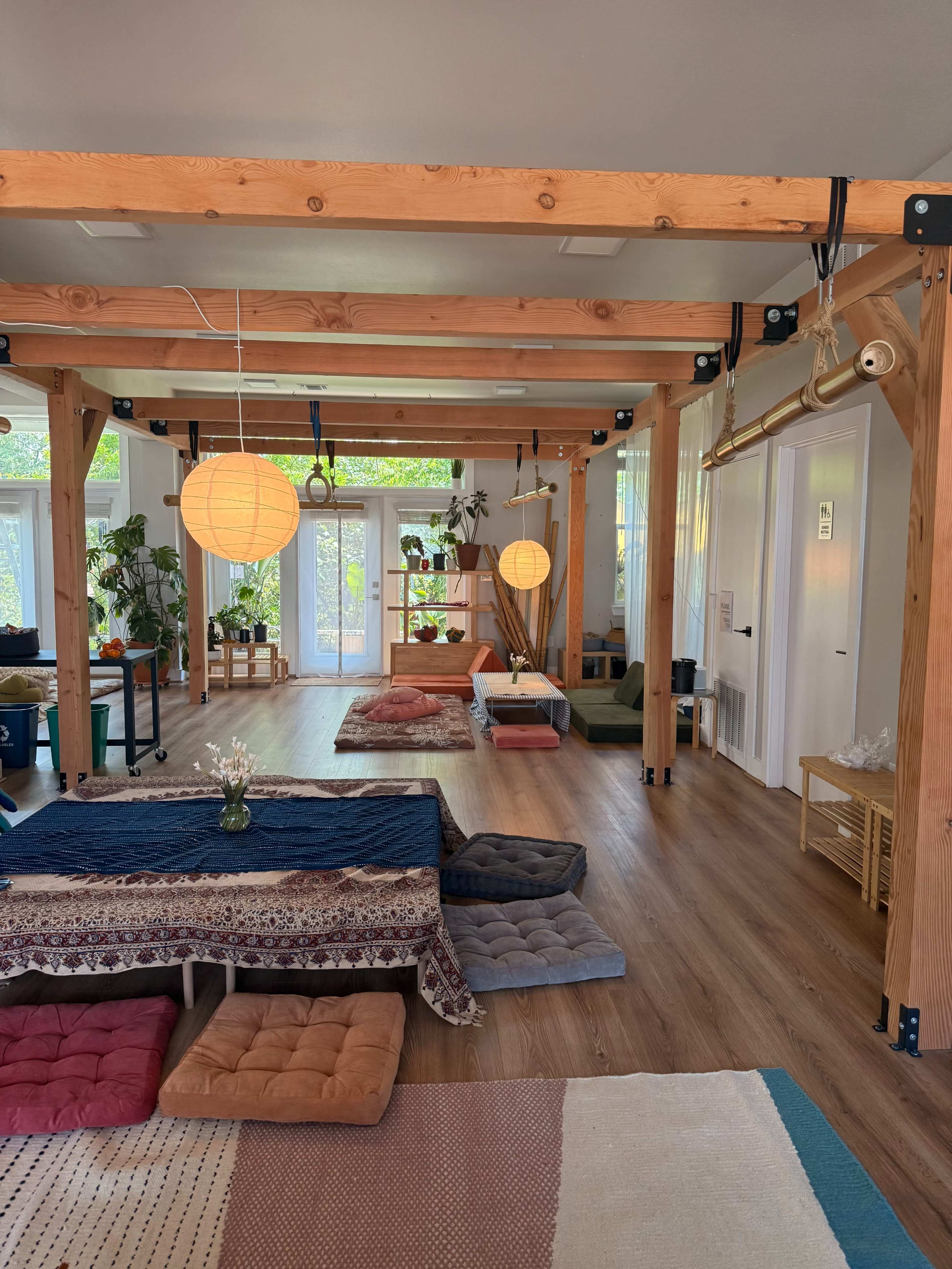 The image shows a spacious room with wooden beams, various floor cushions, and a few indoor plants near large windows allowing natural light.