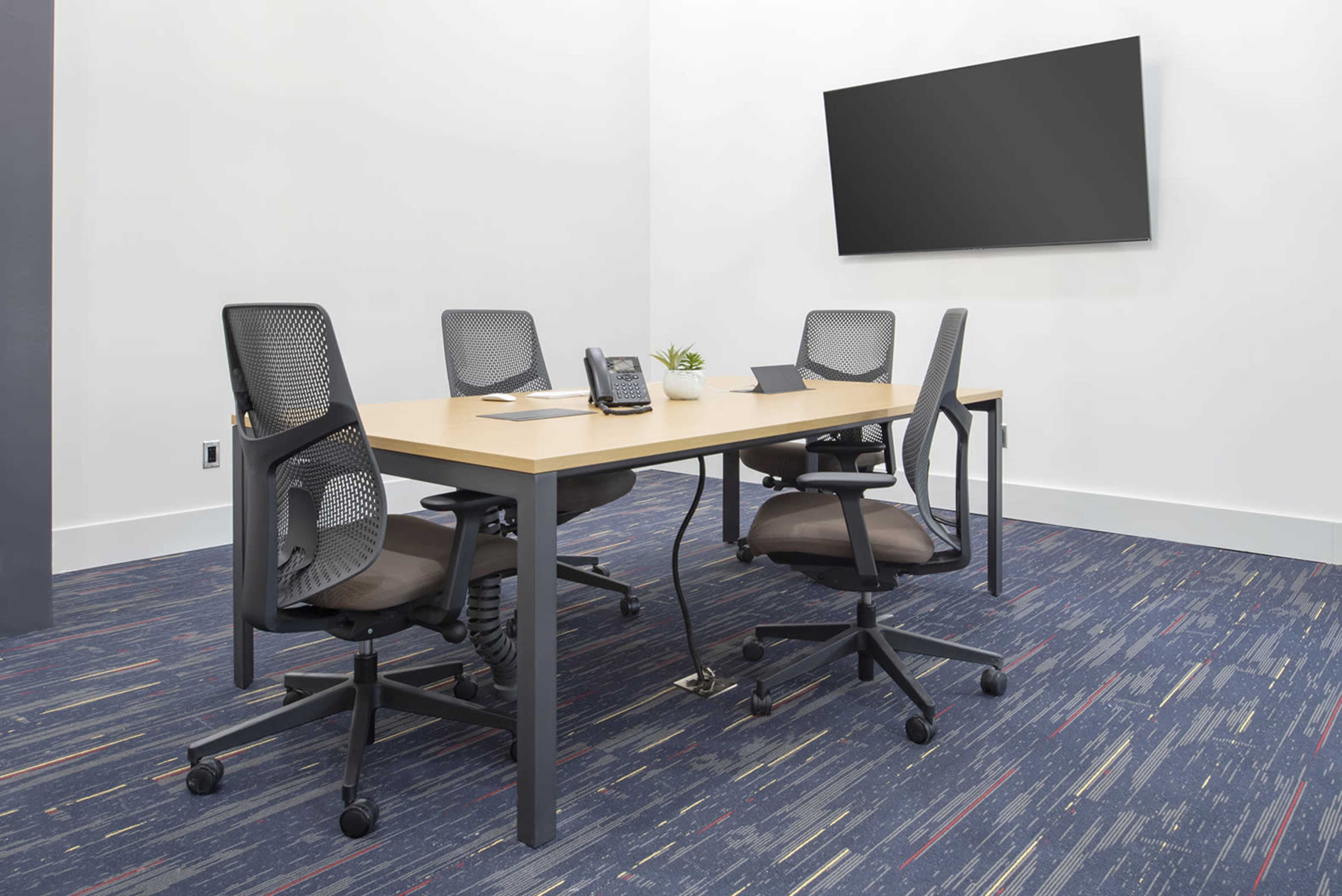 A conference room features a wooden table with four adjustable chairs, a telephone, a laptop, and a blank wall-mounted screen.