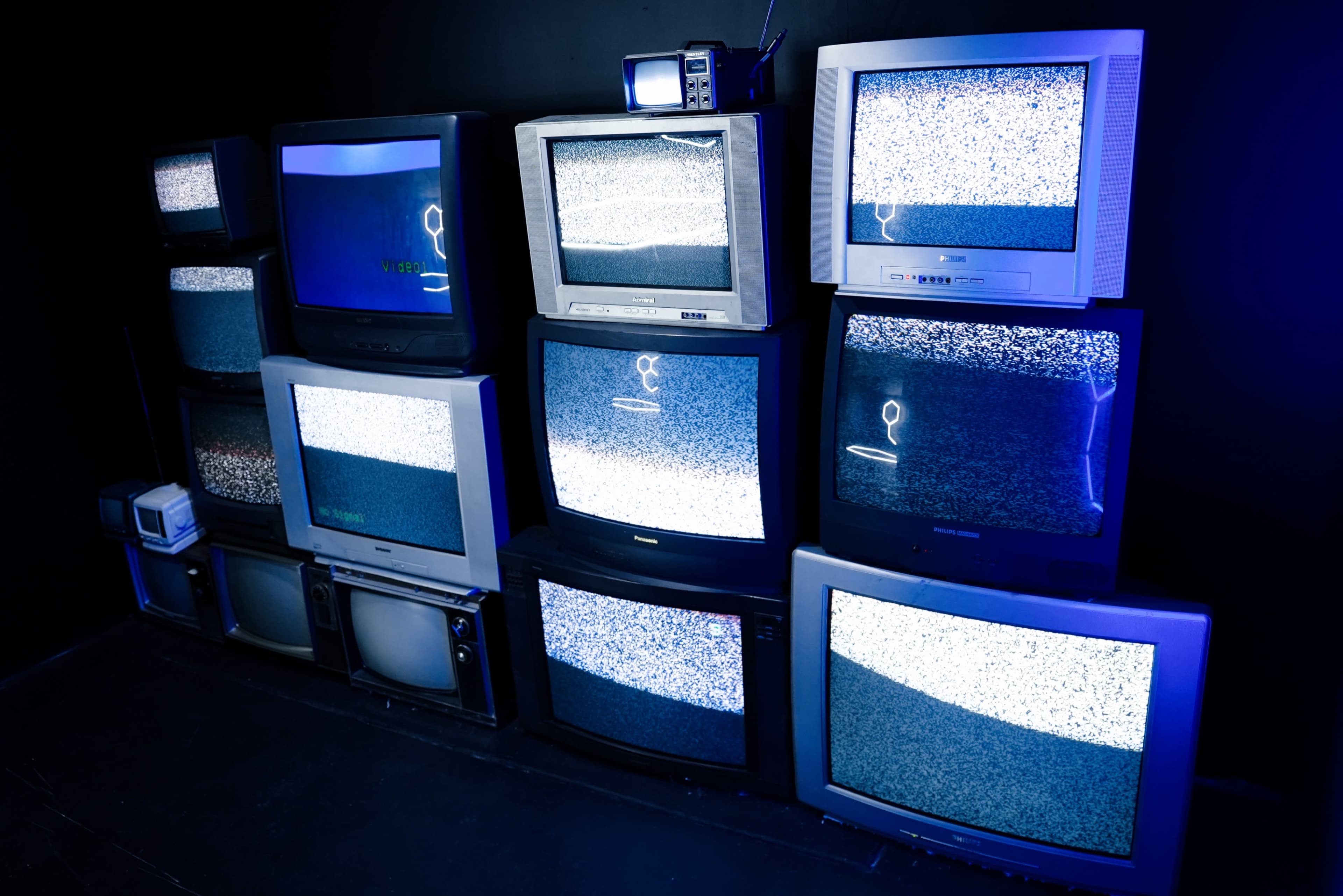 A stack of old television sets displaying static is arranged against a dark wall.